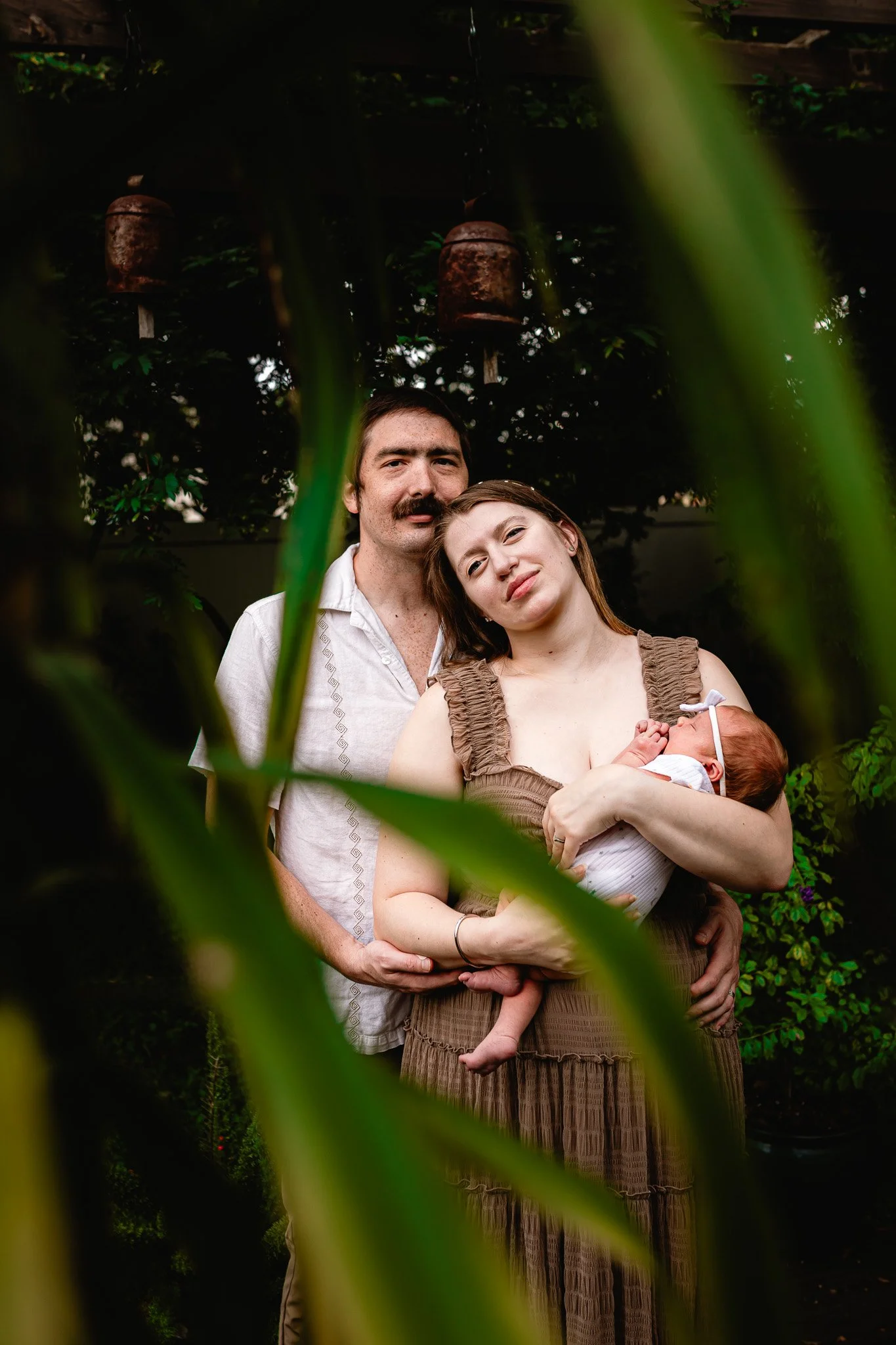A man with dark hair and a mustache, a woman with light brown hair, and a baby in a white outfit are standing together outdoors, partially obscured by green plants in the foreground. The woman is holding the baby, and they are in front of a dark leafy background with hanging rusted bells.