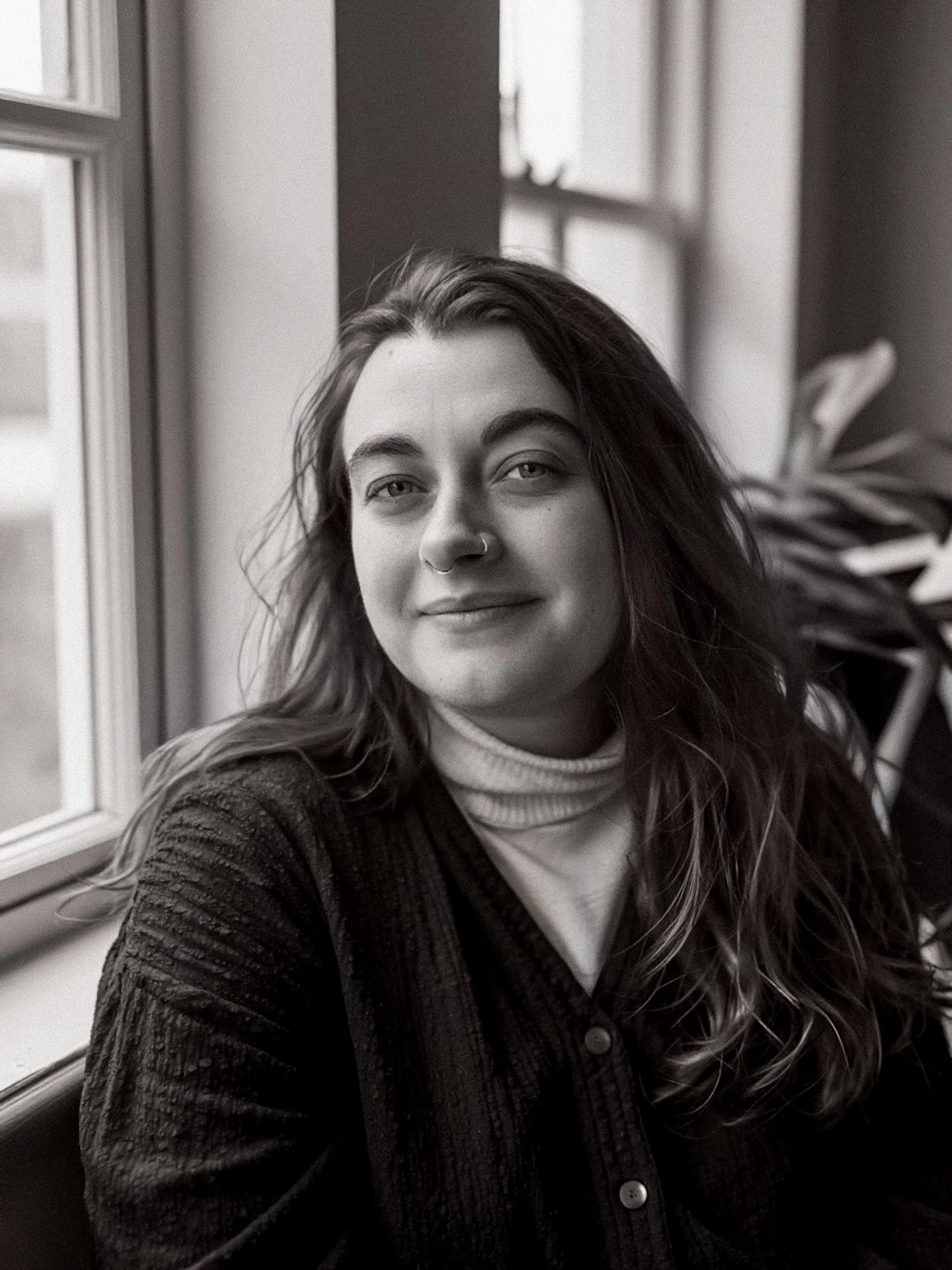 A black and white photo of a woman with long wavy hair, wearing a knitted cardigan over a turtleneck sweater, sitting near a window with a plant nearby.