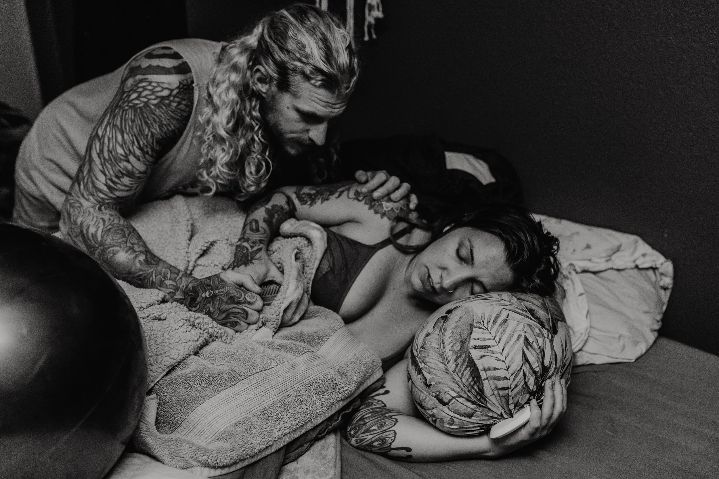 A black-and-white photo of a man with long curly hair and tattoos on his arms, lying on a bed with a woman who has short hair and tattoos on her arms. The man is leaning over the woman, looking at her as she sleeps, covering her with a blanket.