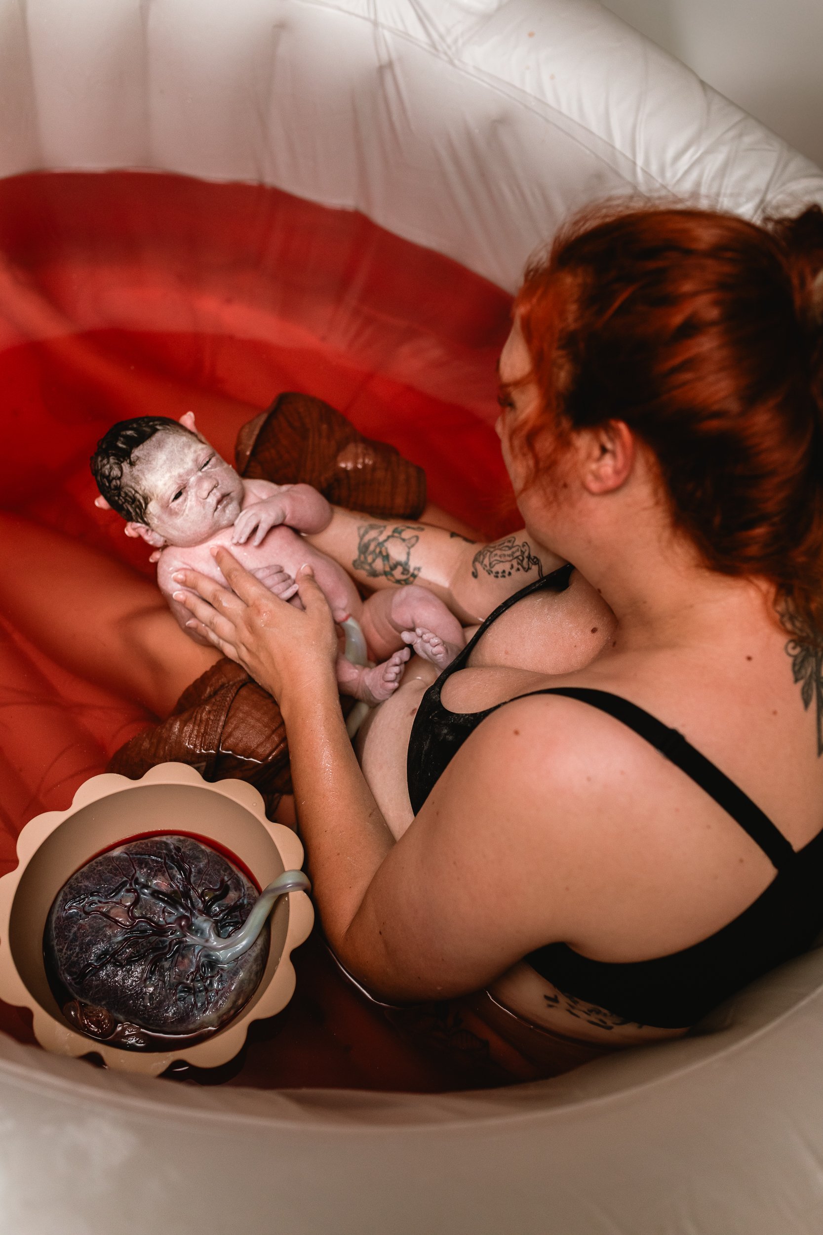 A woman giving birth in a birthing tub, holding a newborn baby with a cord attached, with water and a birth ball visible.