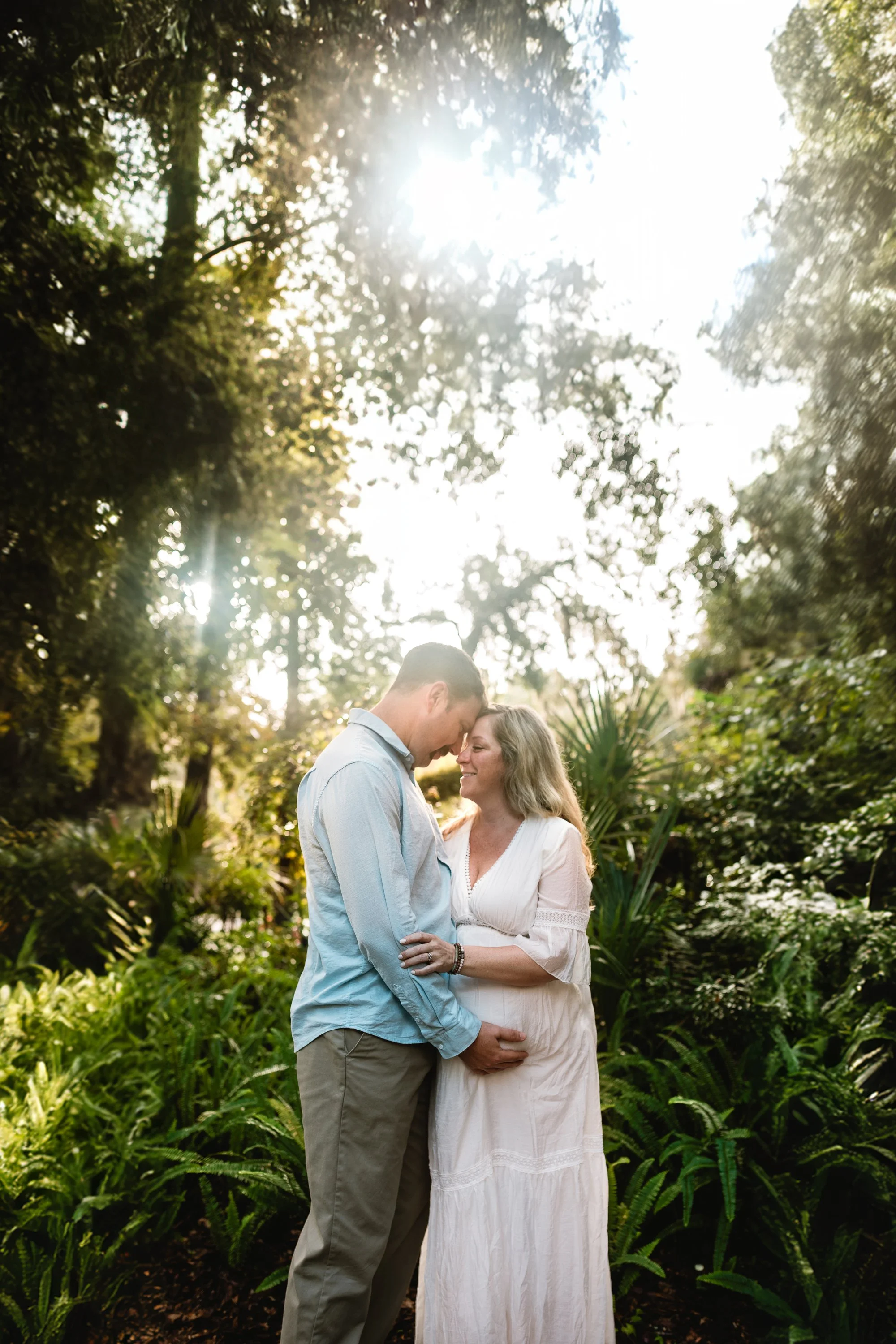 A couple stands close together in a lush, green outdoor setting with tall trees and ferns, touching foreheads and smiling. The sunlight filters through the trees, creating a warm, romantic atmosphere.