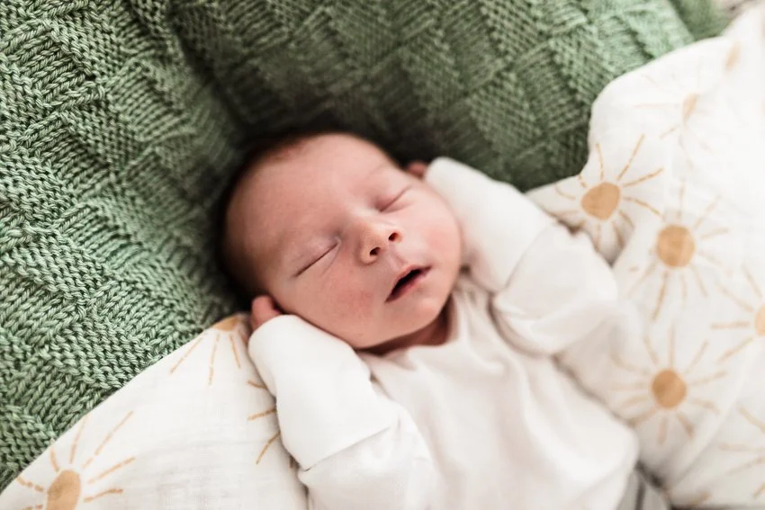 A newborn baby sleeping peacefully on a green knitted blanket with a white blanket decorated with yellow sun patterns partially covering them.