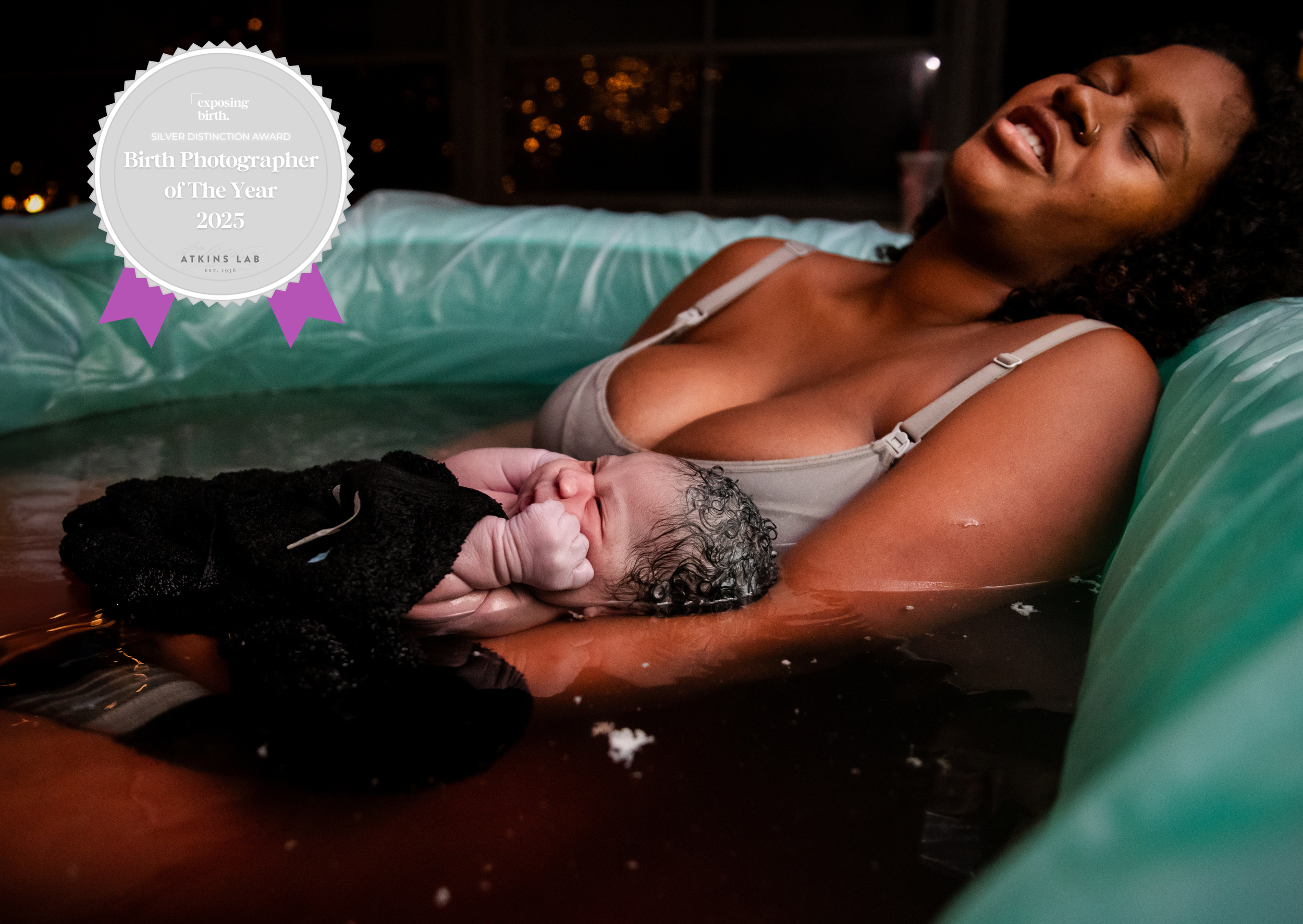 A woman with curly hair and a tan complexion lies in a birth pool, smiling with eyes closed, holding a newborn baby with wet, curly hair on her chest, during a water birth at night.