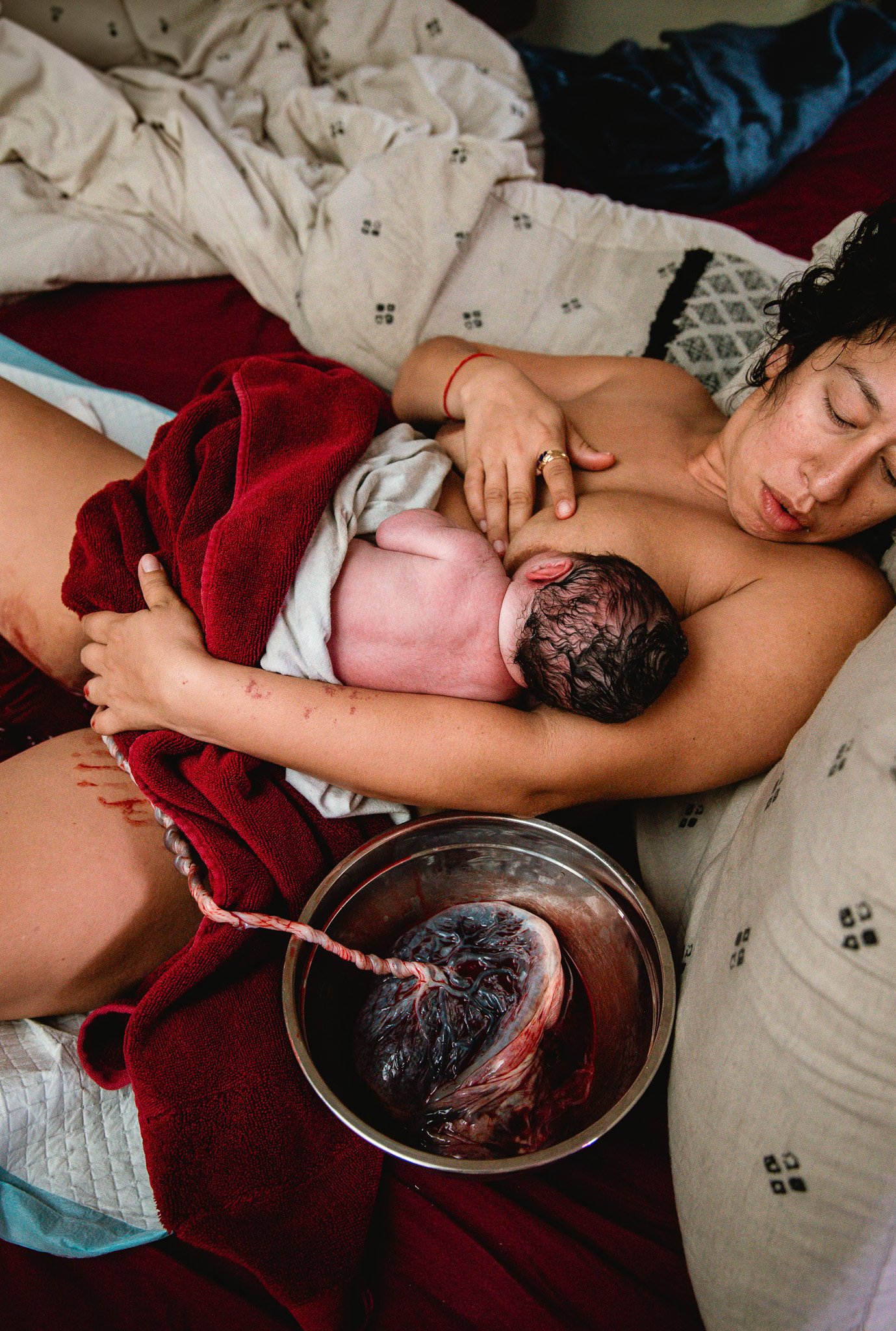 A woman with curly hair is lying in bed with her eyes closed, holding a newborn baby on her chest. There is a bowl of dark red liquid with some tissue inside next to them, and various blankets and bedsheets with different patterns around.