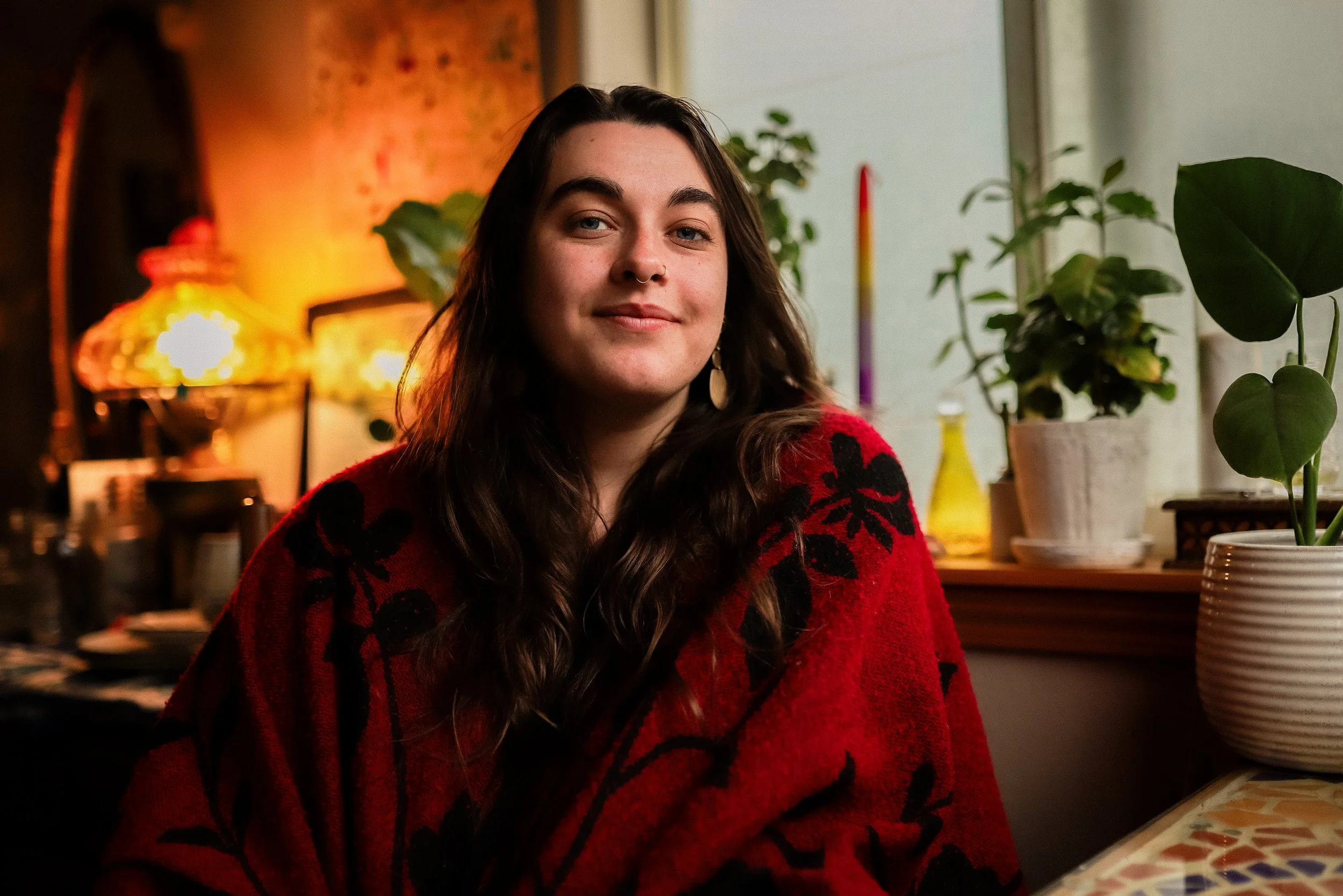 A young woman with long dark hair, wearing a red sweater with black floral patterns, sitting indoors near a window with potted plants behind her, in a warmly lit room.