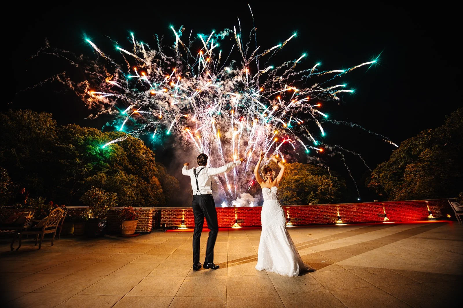 Een bruid en bruidegom vieren hun huwelijk met vuurwerk op een terras 's nachts, omgeven door bomen.