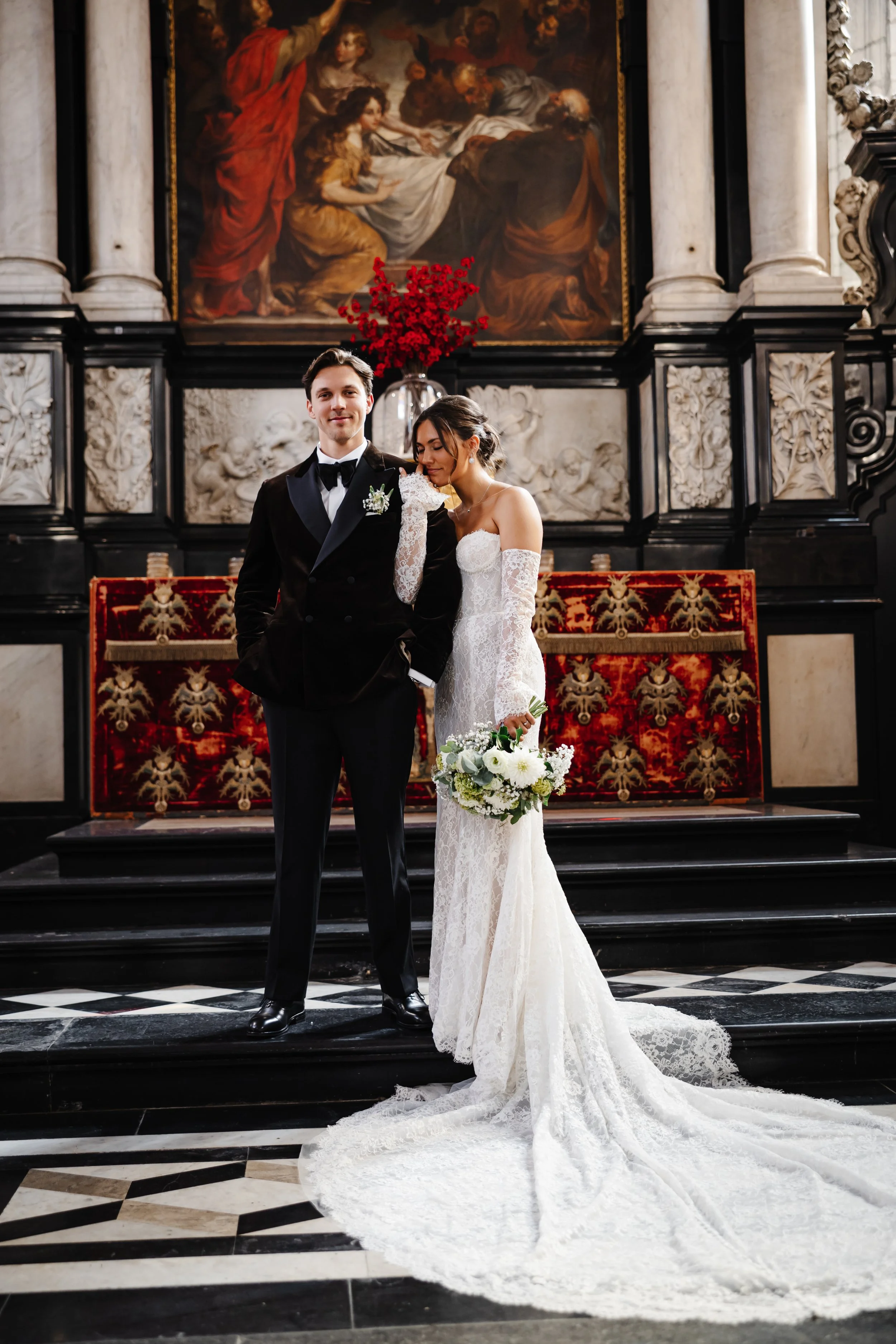 Een getrouwd koppel poseert voor een schilderachtig decor in een kerk. De man draagt een zwarte tuxedo en de vrouw een witte kanten trouwjurk met lange mouwen, en houdt een bloemboeket in haar hand. De achtergrond bevat een groot historisch schilderi