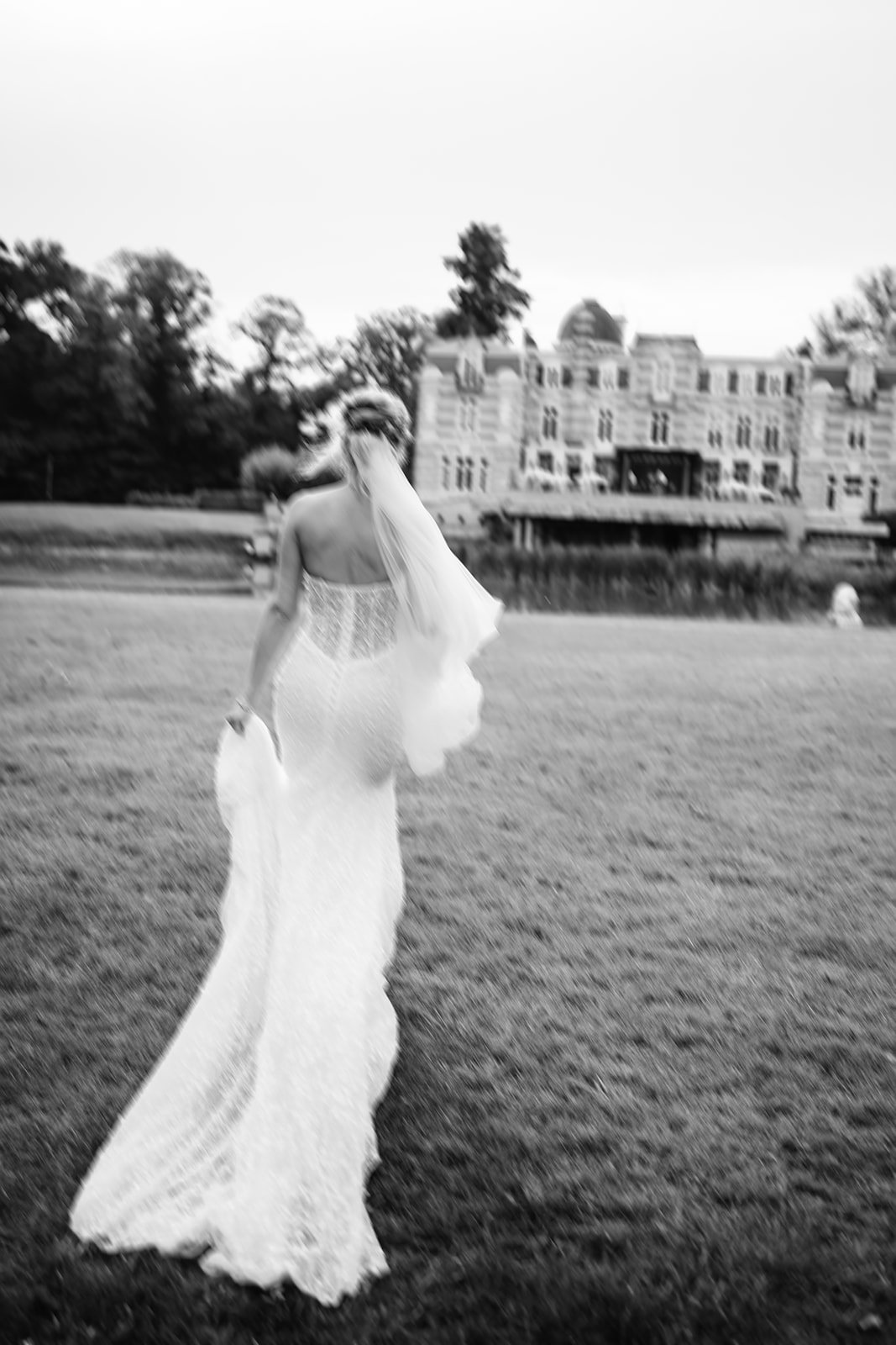 Een vrouw in een witte trouwjurk die door een grasveld loopt, gezien vanaf de achterkant, met een groot herenhuis op de achtergrond in zwart-wand.