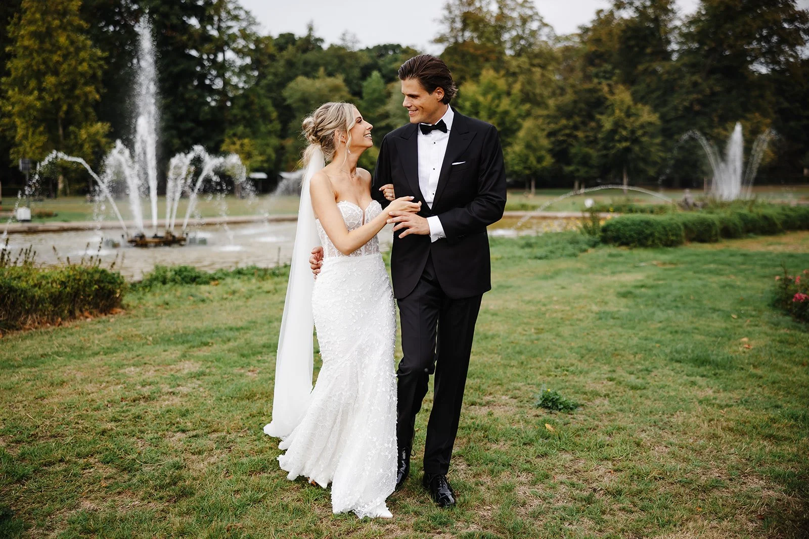 Bruid en bruidegom dansen in een park met fonteinen op de achtergrond.
