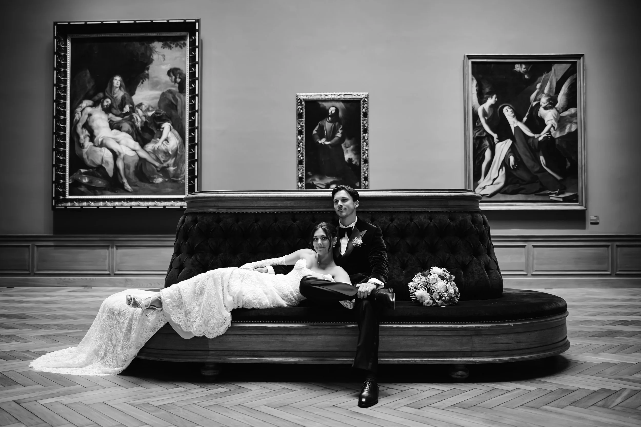 Een zwart-wit foto van een getrouwd stel dat op een bank in een kunstmuseum ligt, omringd door klassieke schilderijen.