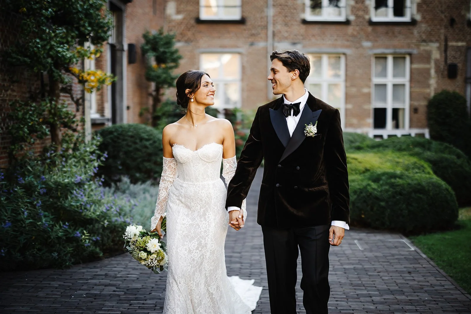 Een bruid en bruidegom lopen hand in hand op een pad in een tuin, glimlachend naar elkaar.