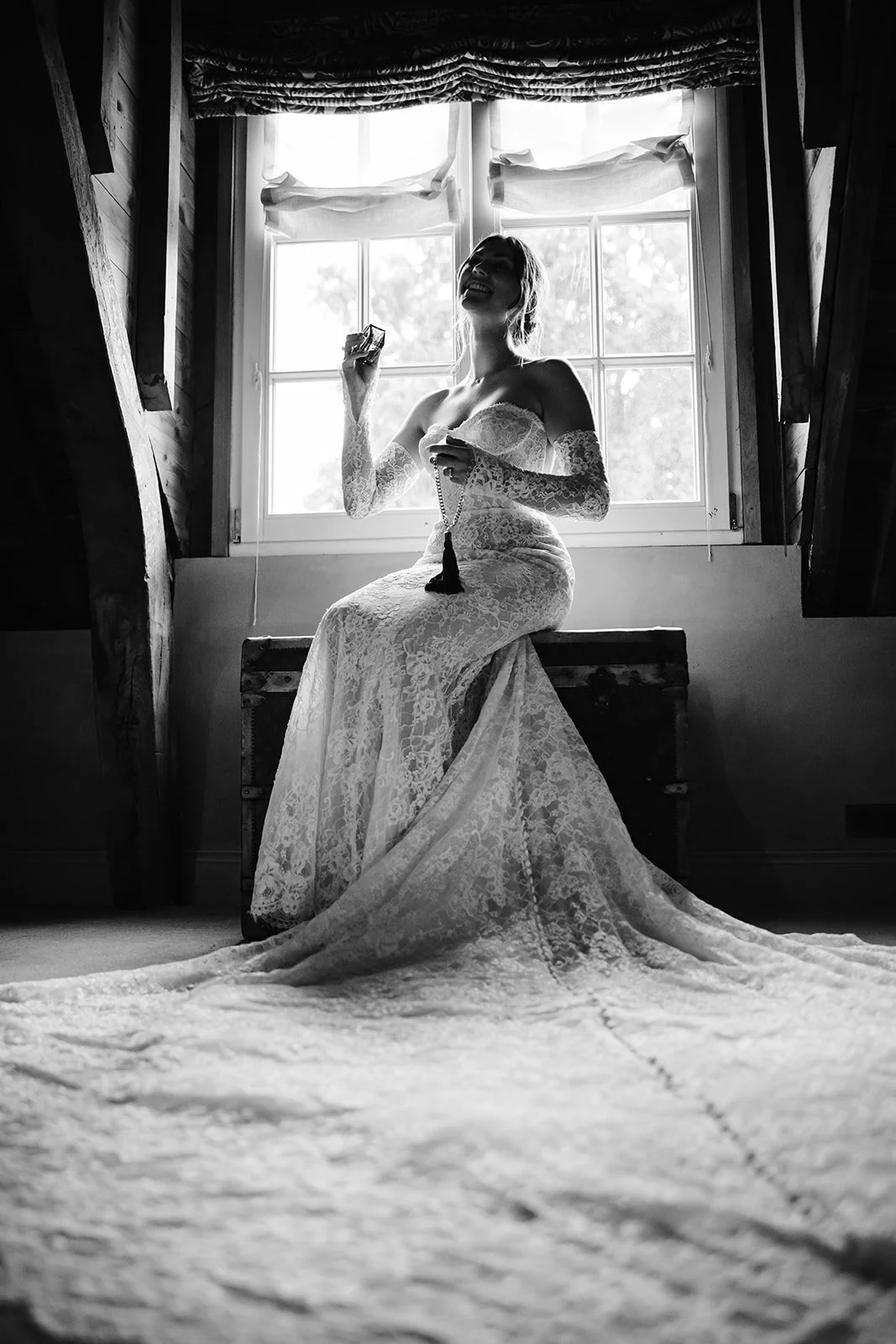 Zwart-wit foto van een vrouw in een kanten trouwjurk zittend op een houten kist voor een raam in een houten kamer, lachend en met een ketting in haar hand.