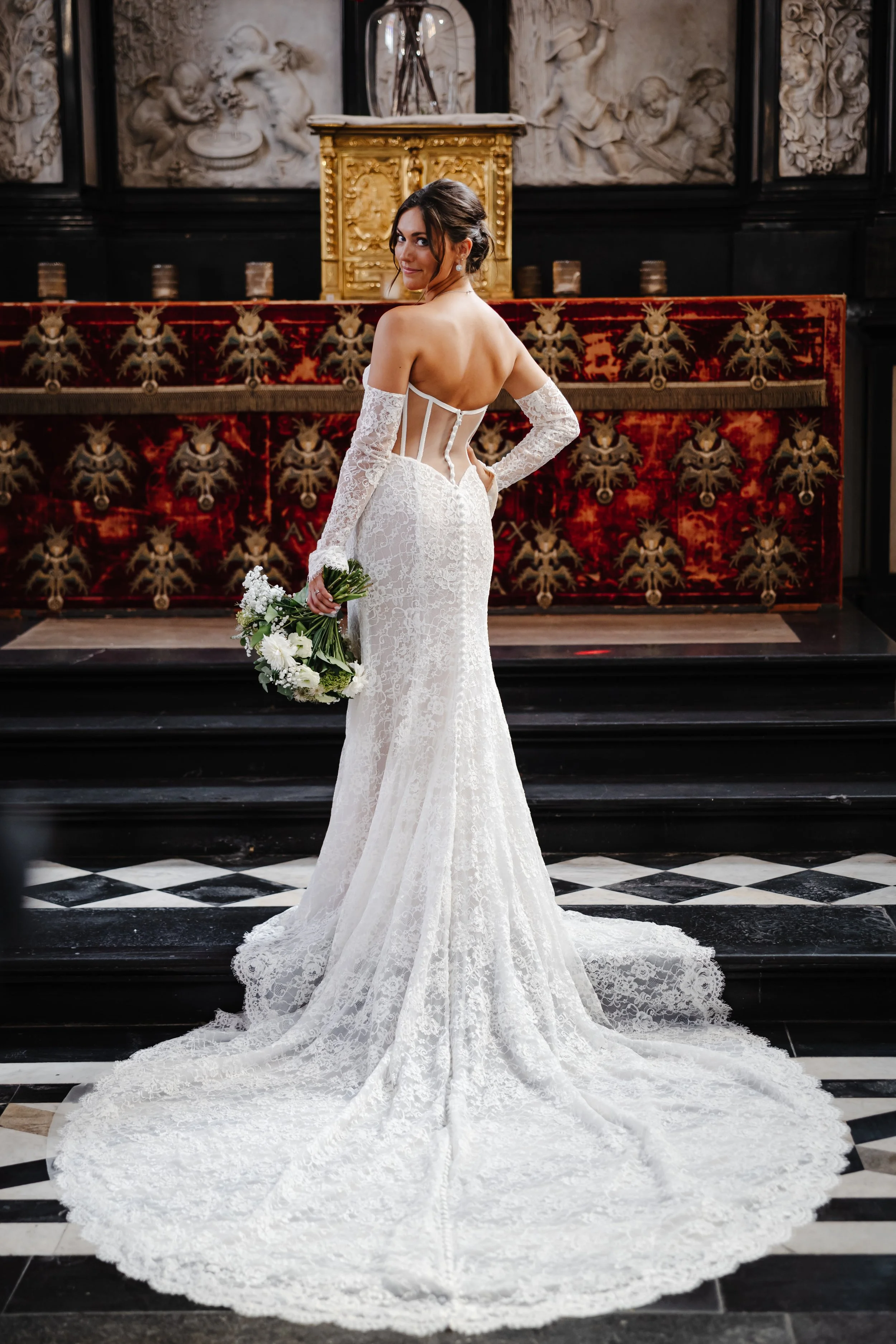 Vrouw in een witte kanten trouwjurk met lange mouwen en open rug, standing in een kerk, die een bloemboeket vasthoudt.
