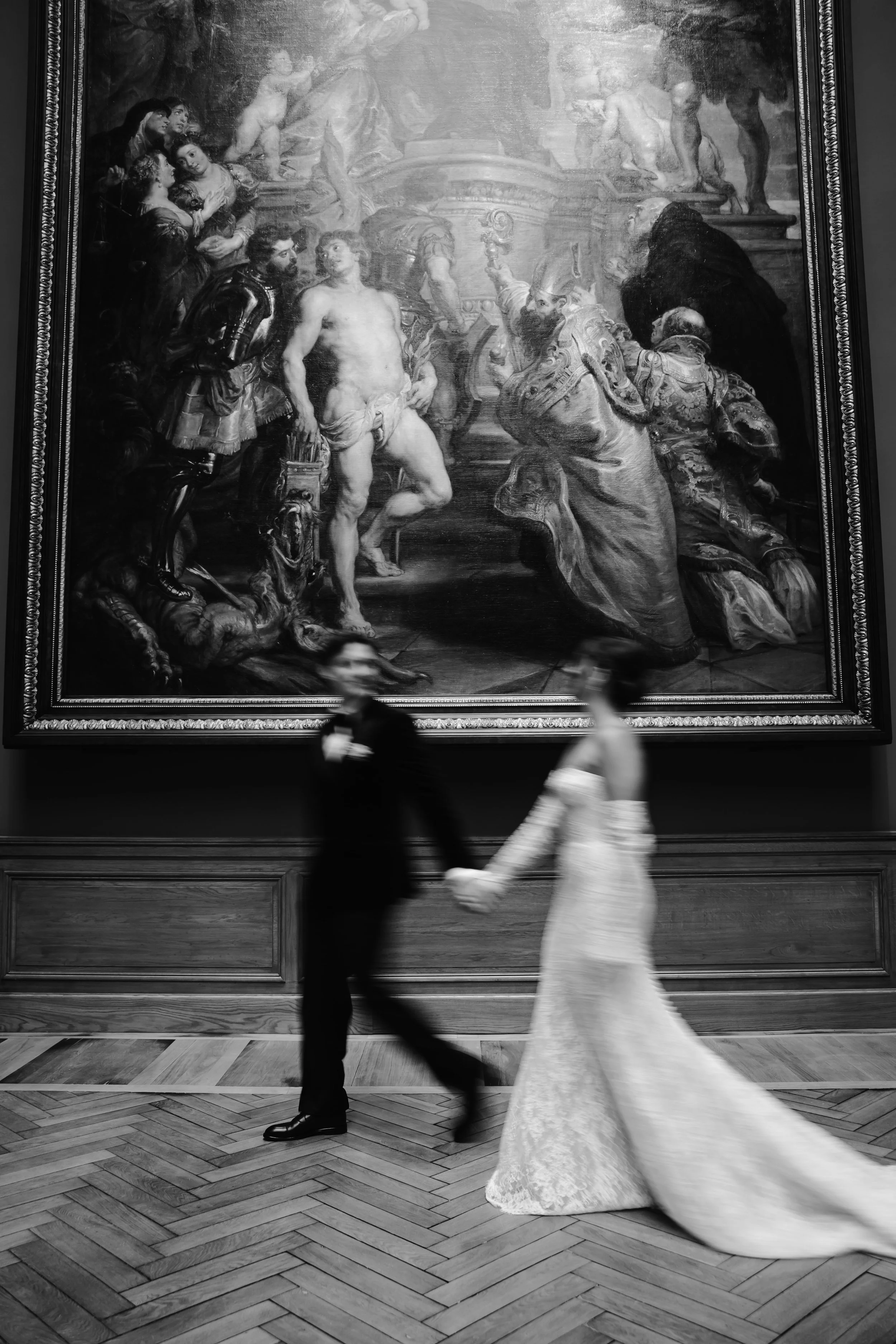 Een koppel in trouwkleding dat door een kunstmuseum loopt, met een grote zwarte en witte muurschildering op de achtergrond.