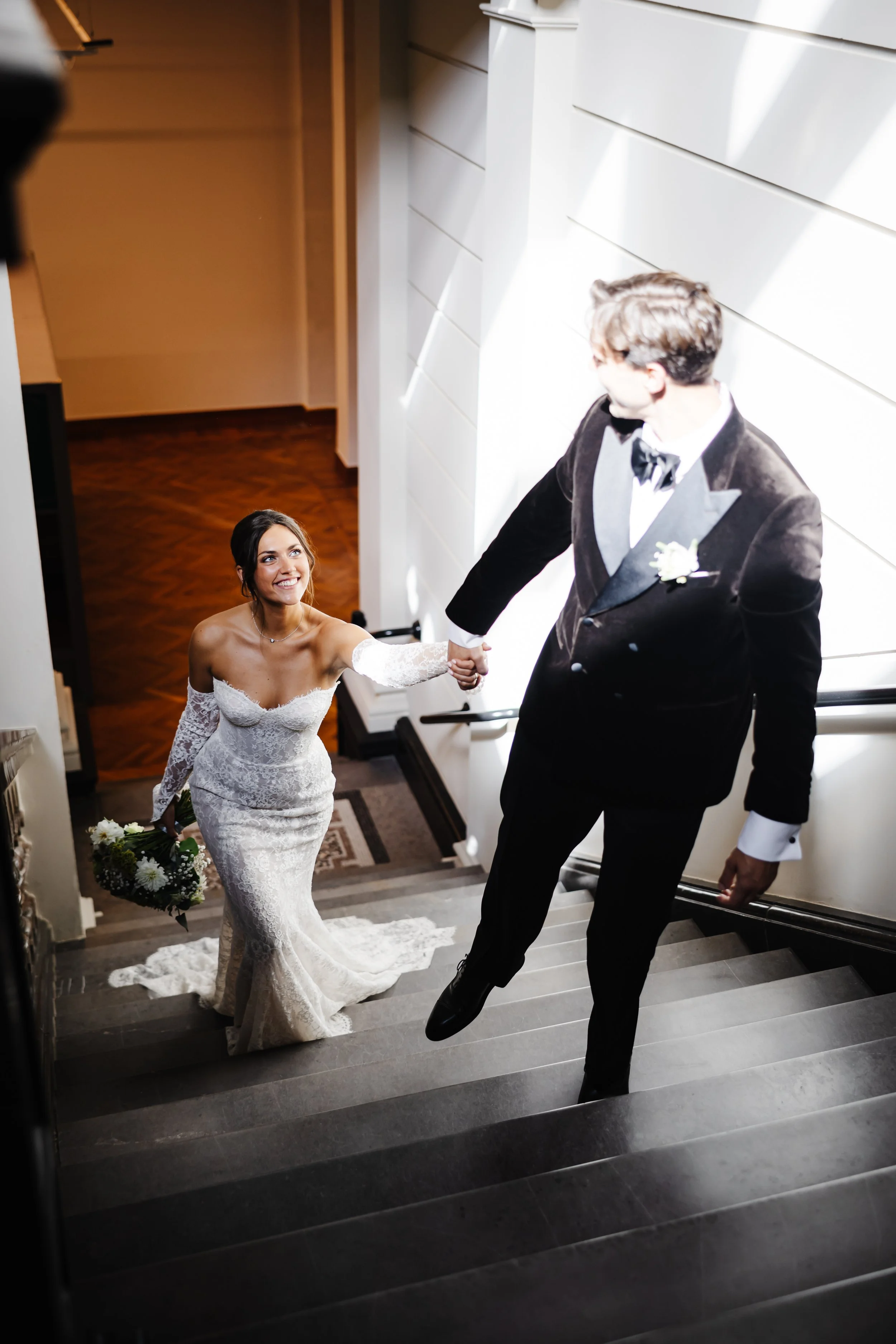 Een bruid en bruidegom die elkaar de hand vasthouden op een trap, zij lacht terwijl hij omhoog komt, zij draagt een witte trouwjurk en hij een zwarte tuxedo.
