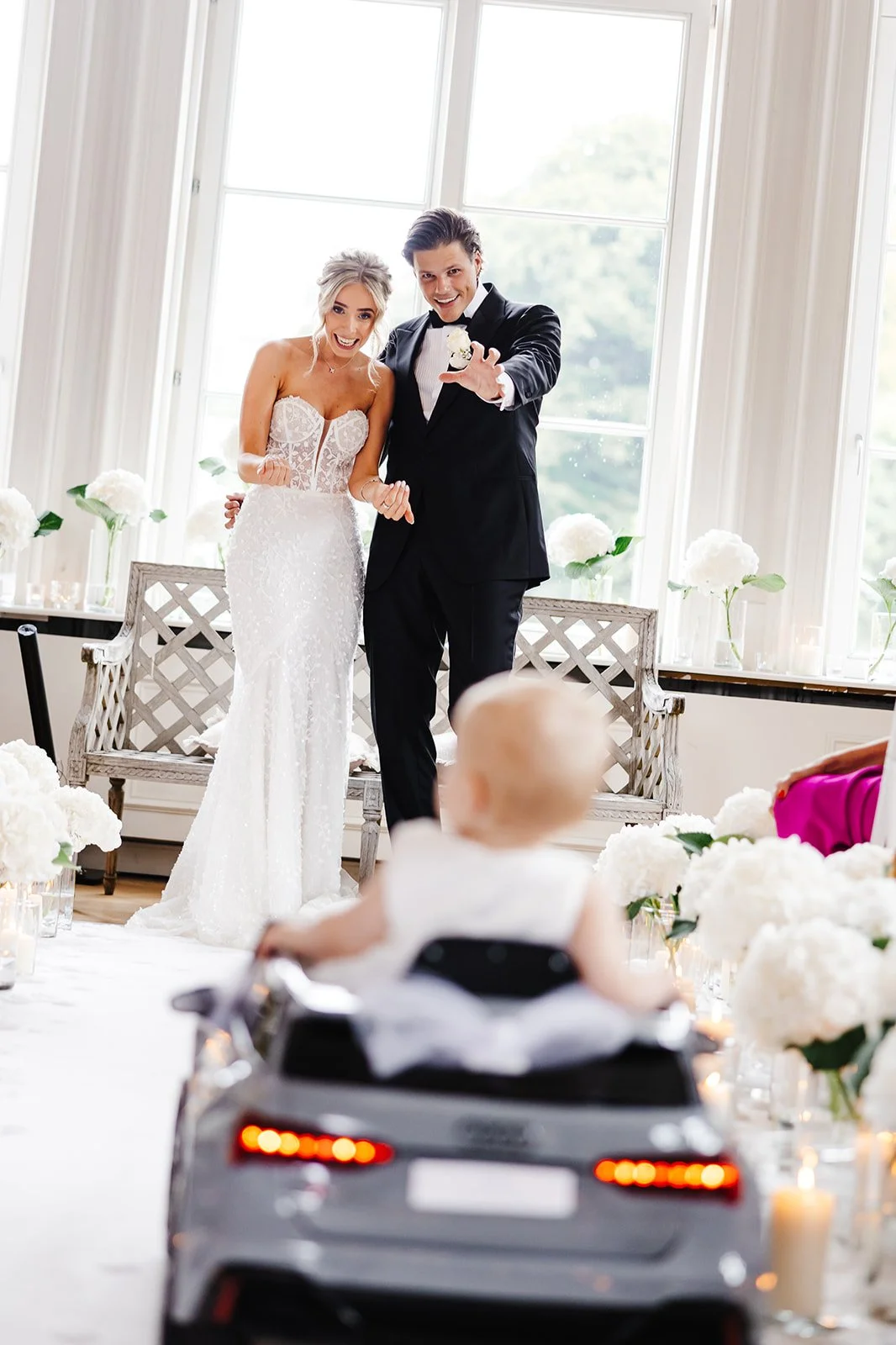 Trouwbriefing met een bruid en bruidegom die op het altaar staan, een kind dat in een speelgoedauto zit kijkt toe, in een lichte, elegante zaal met grote ramen en witte bloemen.