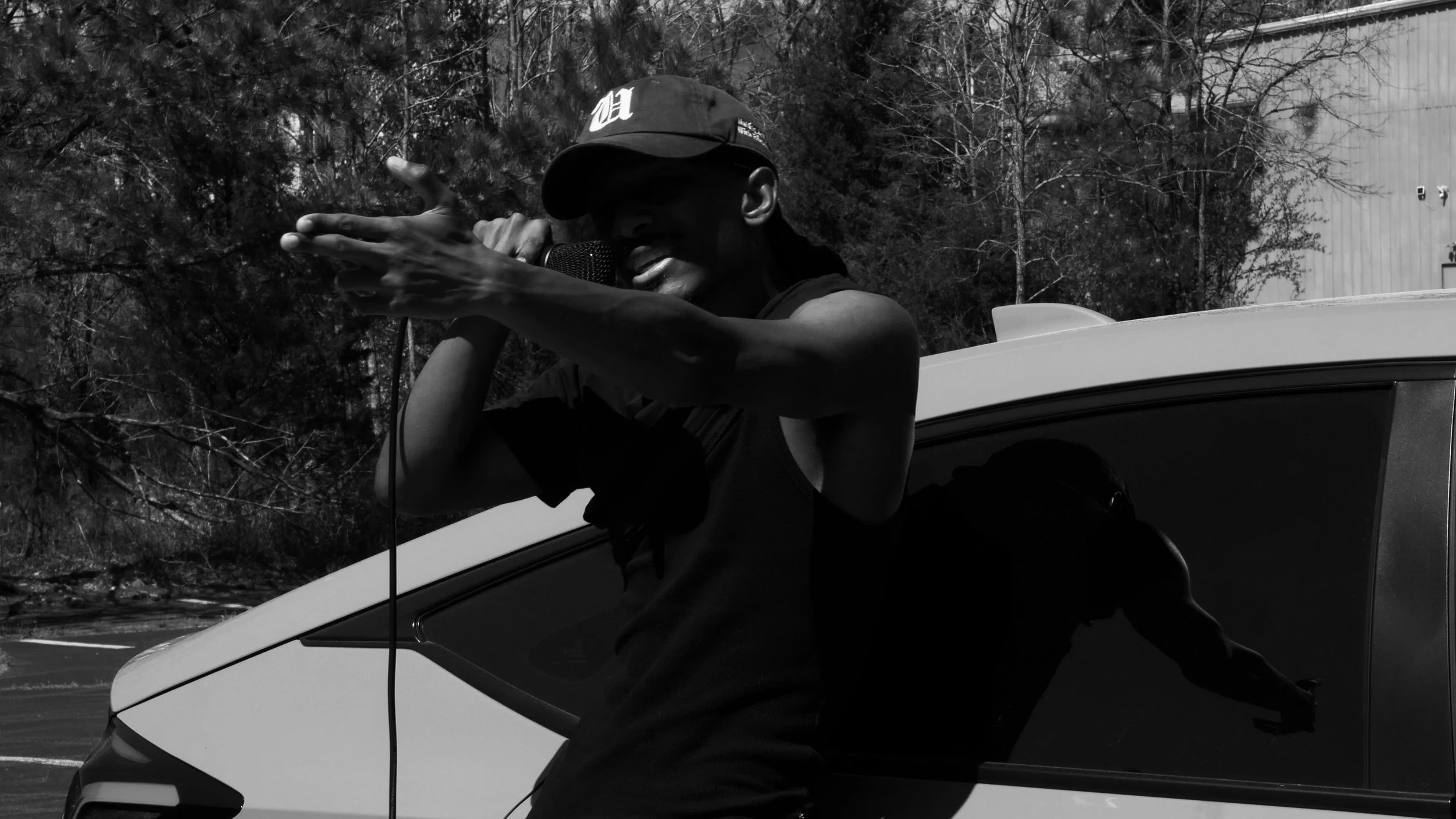 Black and white photo of a person wearing a cap, holding a microphone, and posing with a finger gun gesture in front of a car.