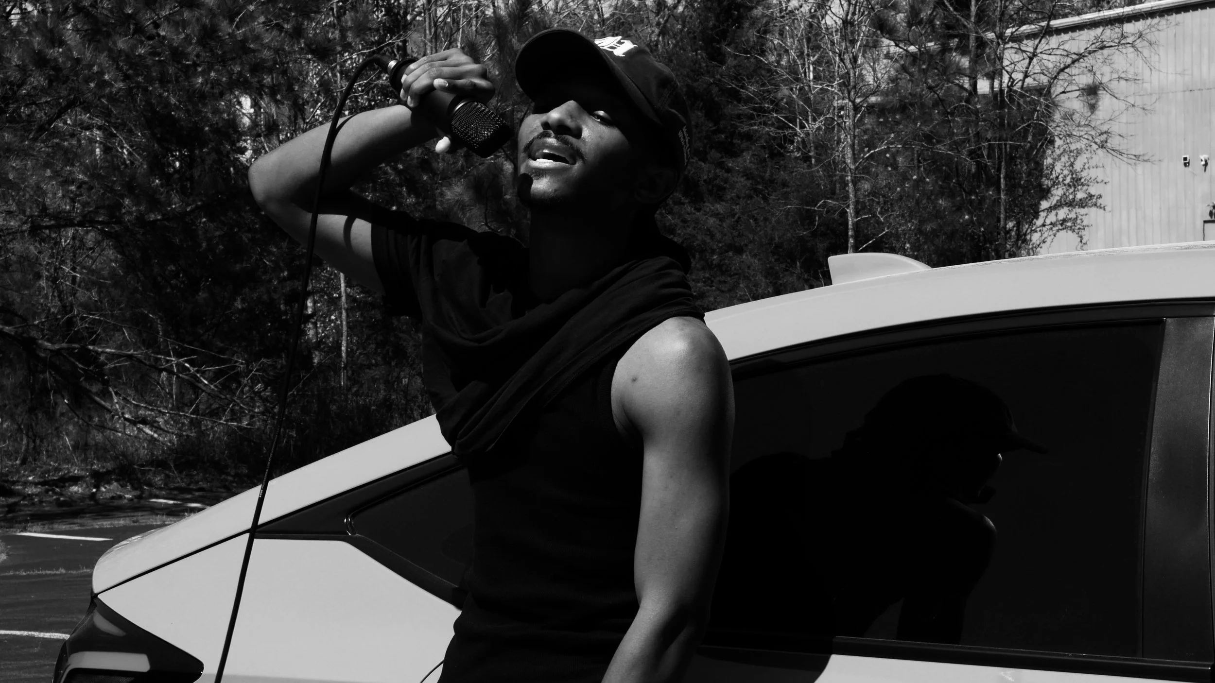 Black and white photo of a person standing by a car, holding a microphone, wearing a cap and sleeveless shirt.