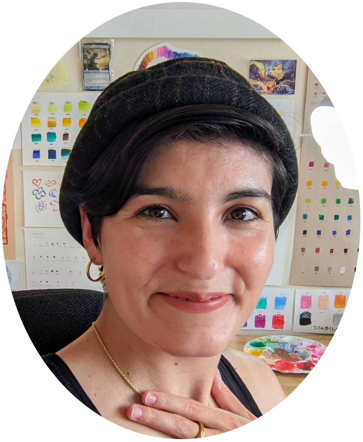 Close-up of a woman with dark eyes and short dark hair wearing a plaid hat, gold jewelry, and a black top. She is smiling directly at the camera. In the background, there are color swatches and artwork.