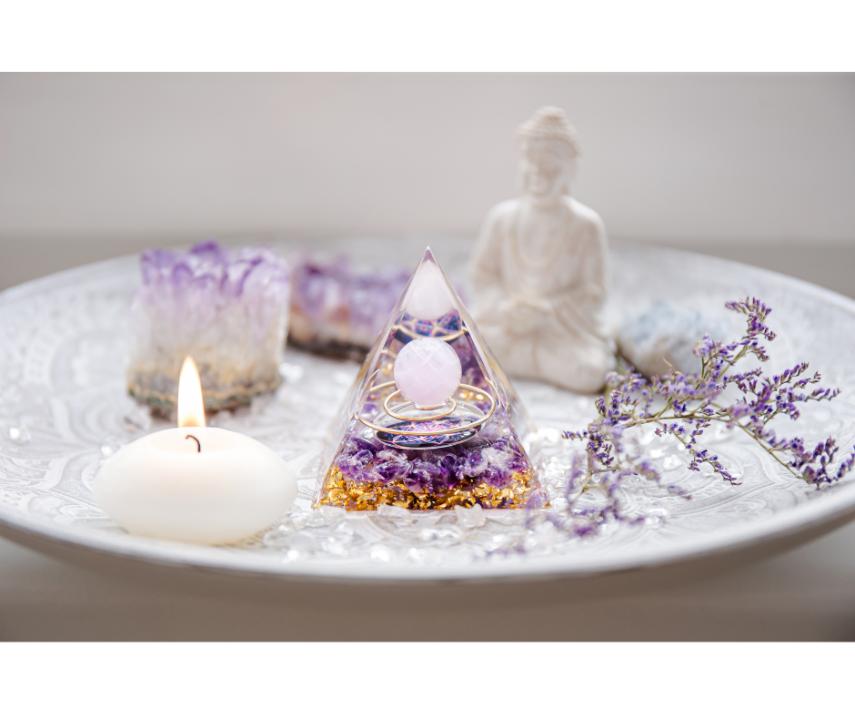 Decorative tray with purple amethyst candle, a white Buddha statue, a purple flower, a glowing candle, and a pyramid-shaped resin ornament with a ring and sphere inside.