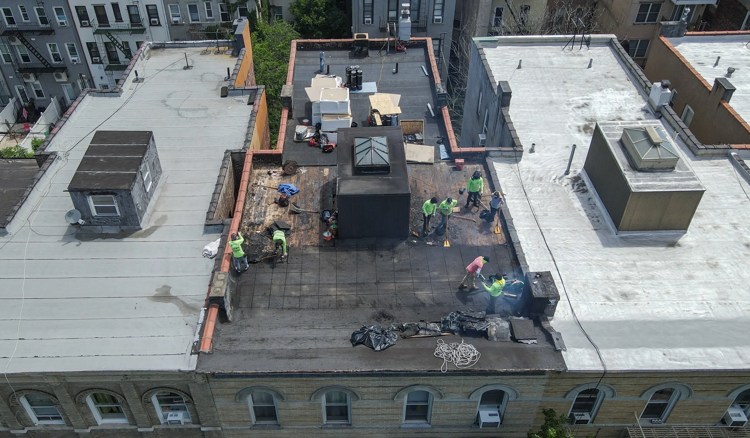 Workers in bright green shirts are repairing and waterproofing the flat rooftop of a building, with some areas covered in black roofing material.