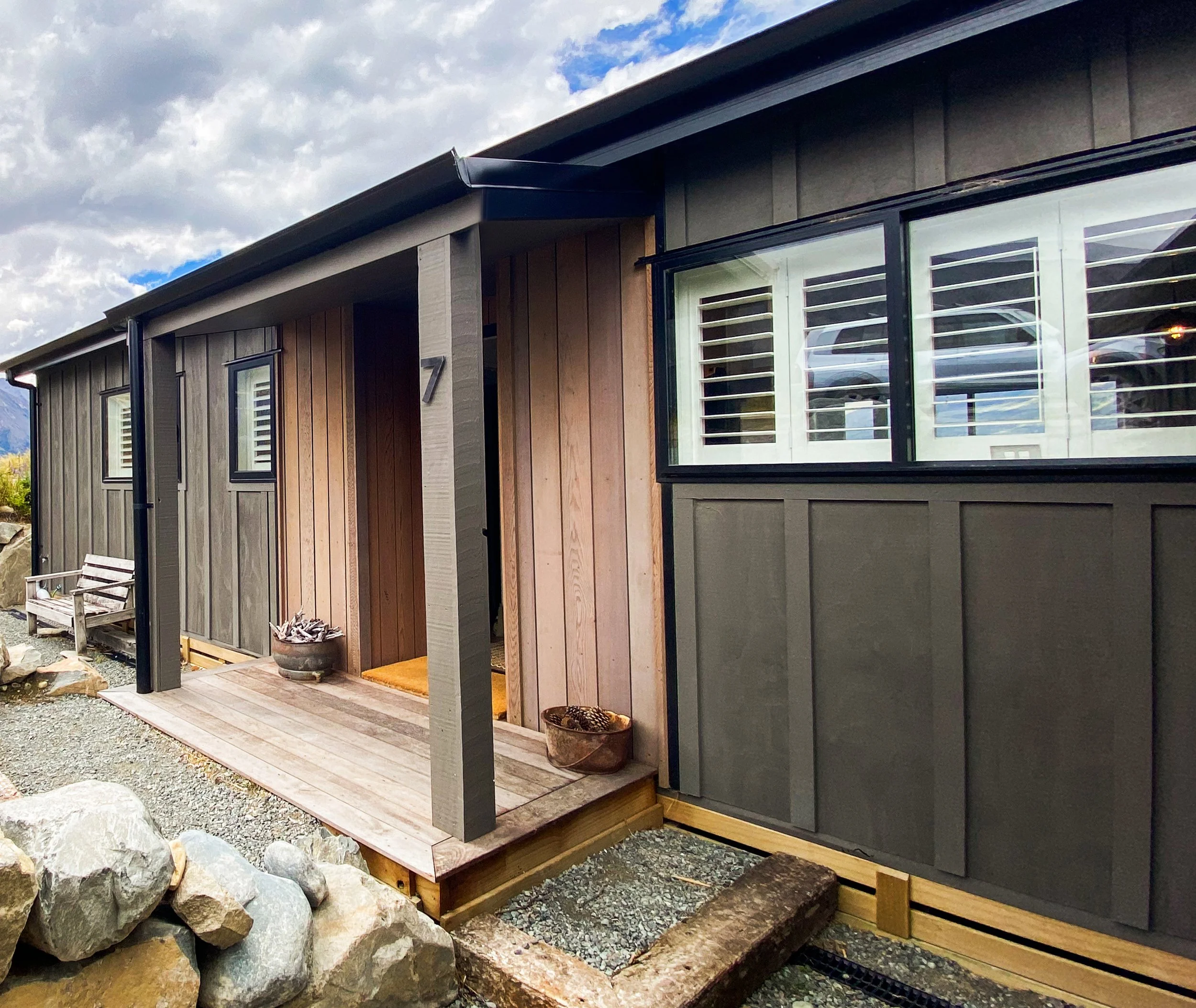Exterior view of a modern house with wooden and gray siding, large windows with white shutters, a small porch with a bench, potted plants, and a gravel pathway surrounded by rocks, under a partly cloudy sky.