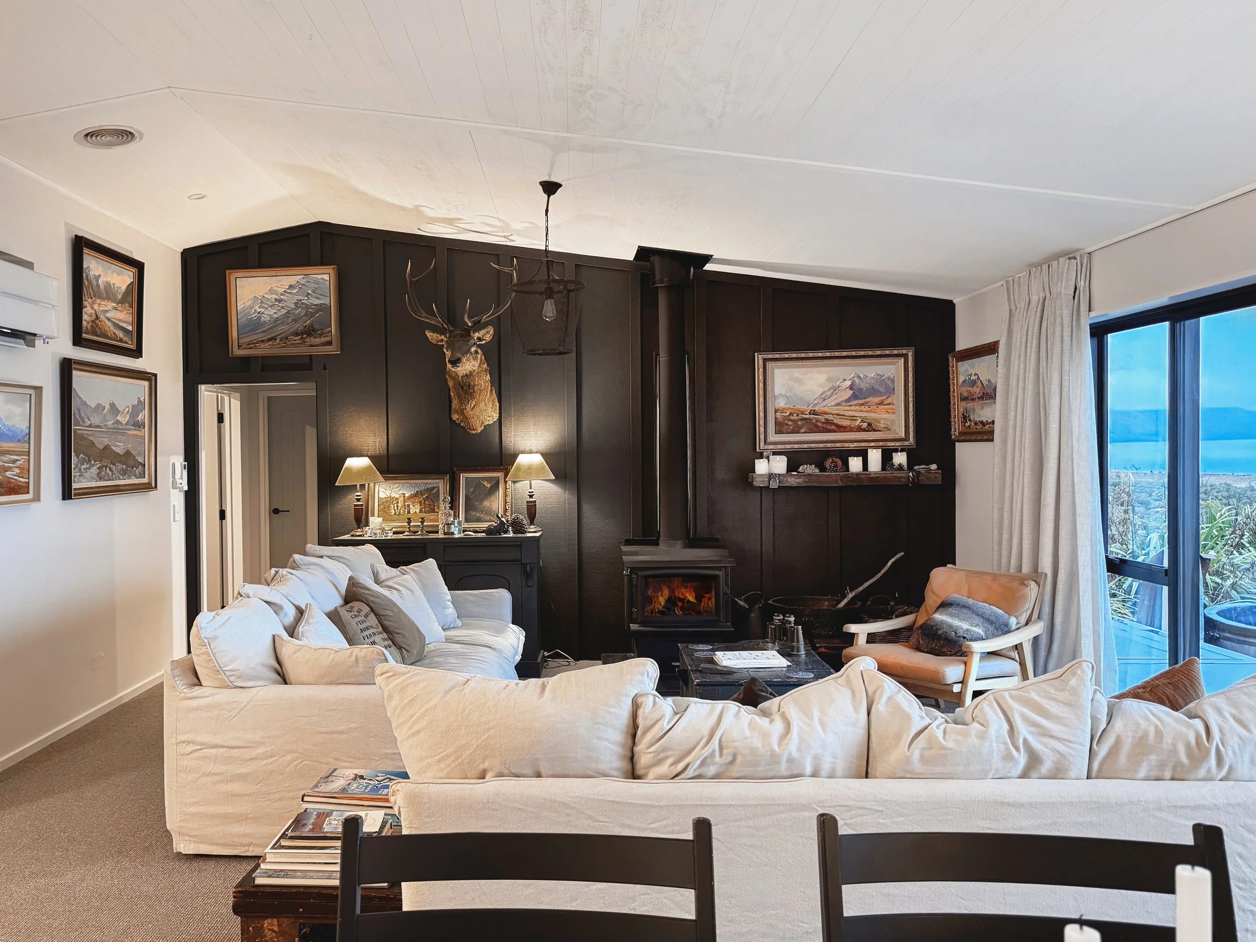 A cozy living room with a large white sofa, a wooden coffee table, and a black accent wall with framed mountain landscape artwork and a mounted deer head with antlers. There are lamps, candles, and decorative items on the black wall. A window with white curtains shows a view of the outdoors.
