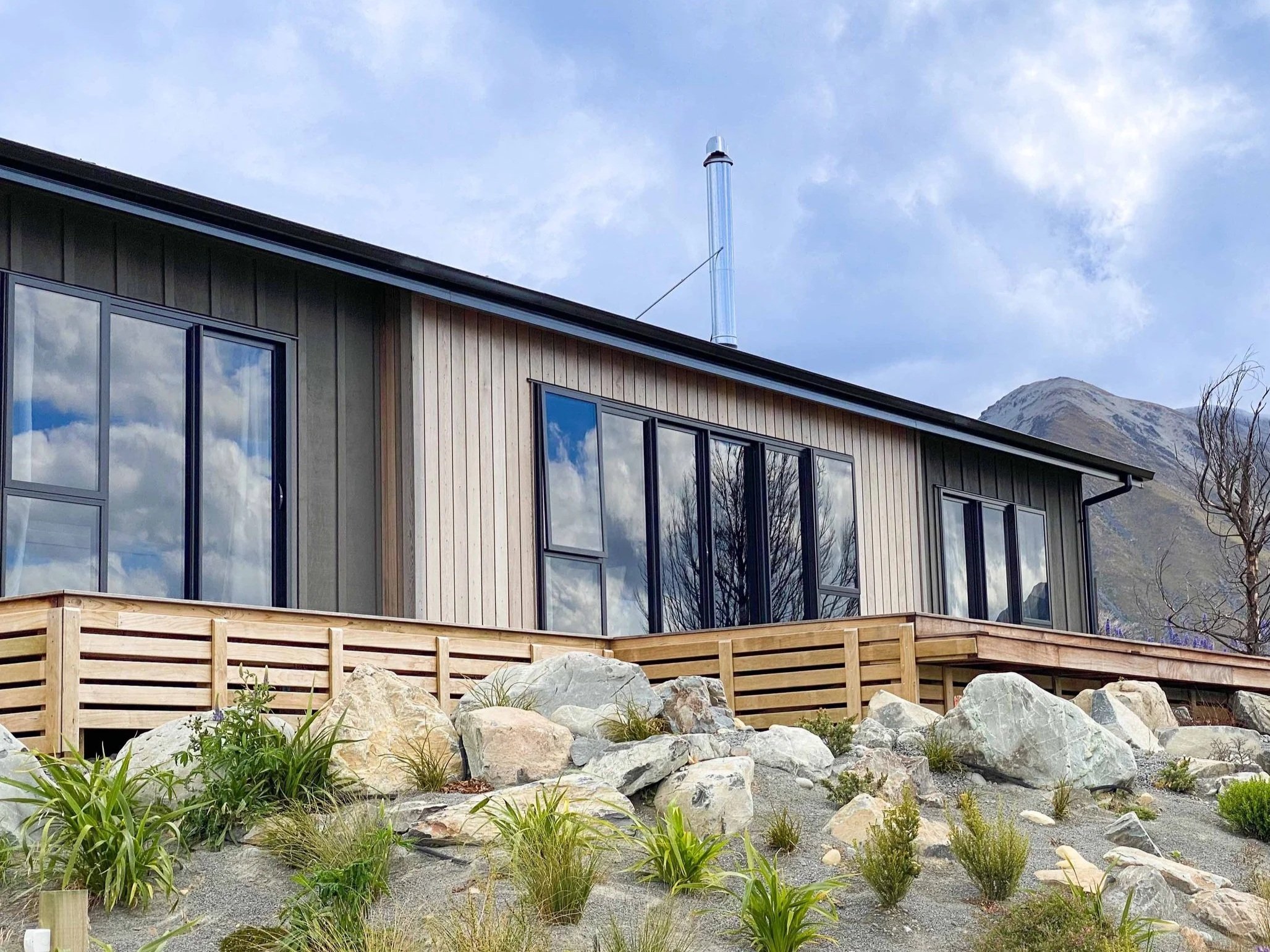 Modern two-story house with large glass windows, wooden siding, and a metal roof, situated in a mountainous area with trees and rocks in the foreground.