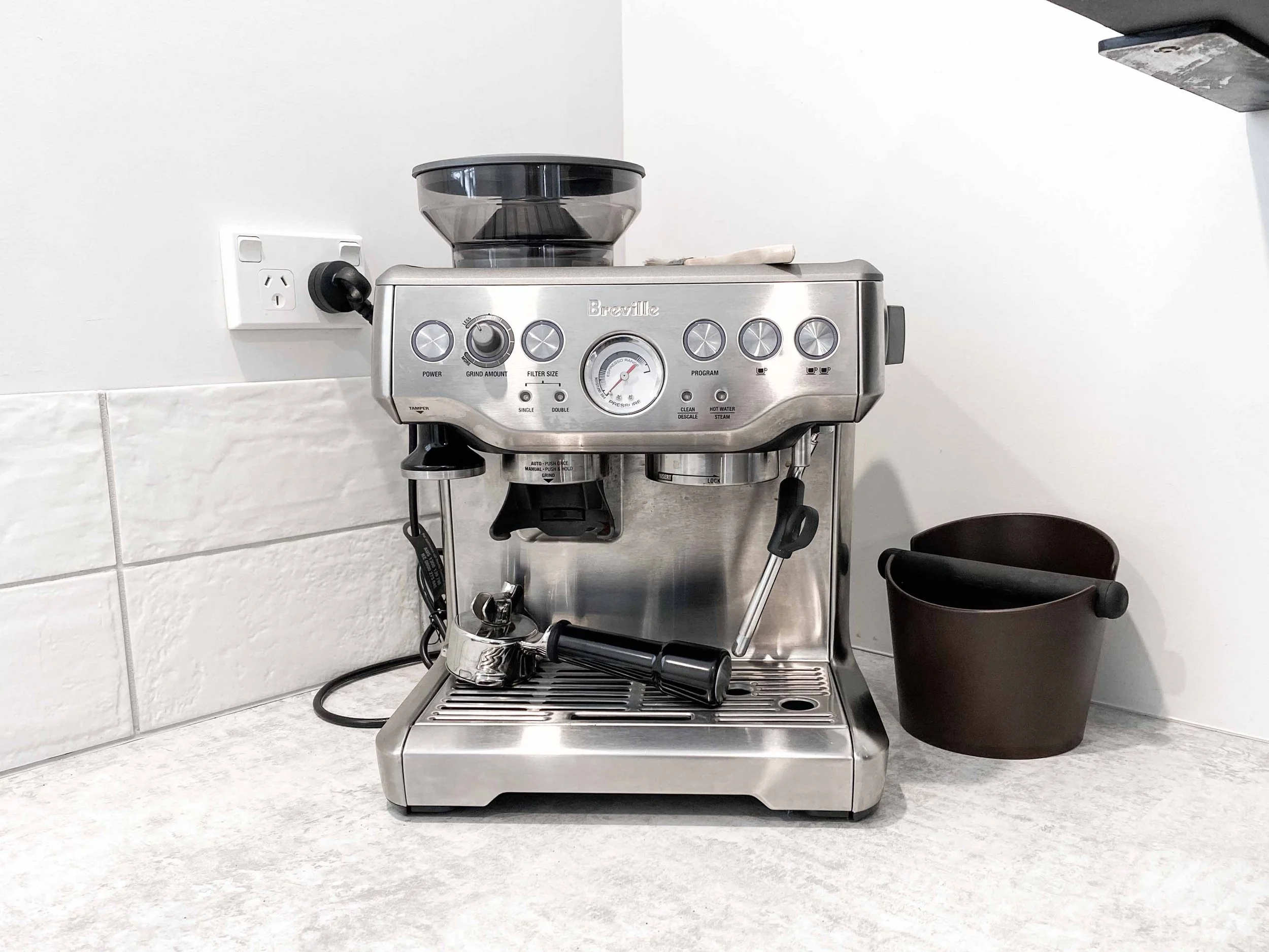 A stainless steel espresso machine with black controls on a white countertop against a white tiled wall.