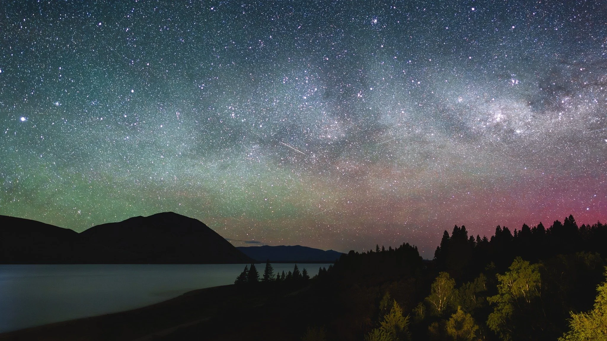 A night sky filled with countless stars, including the Milky Way galaxy, over a landscape of mountains, a body of water, and trees with some pink and purple hues near the horizon.