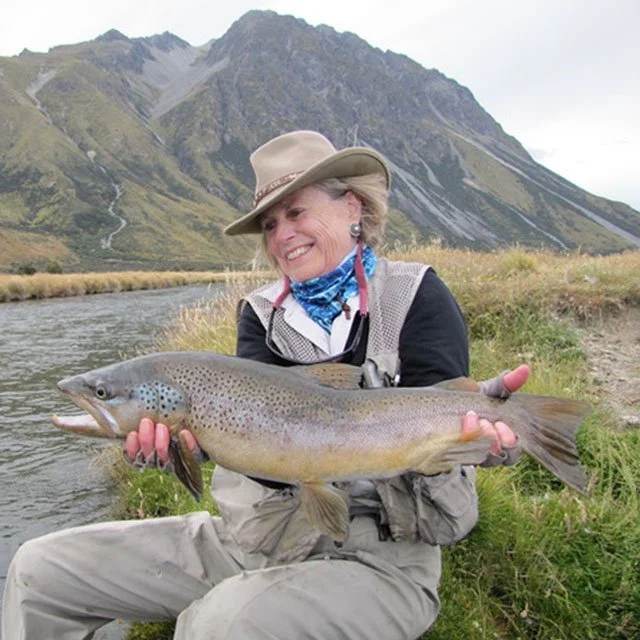Smiling woman in outdoor gear holding a large fish near a river with mountains in the background. Twizel Ohau Canal Lake River Fishing Salmon Trout. Stay at Lake Ohau Escape AirBnb Accomodation.