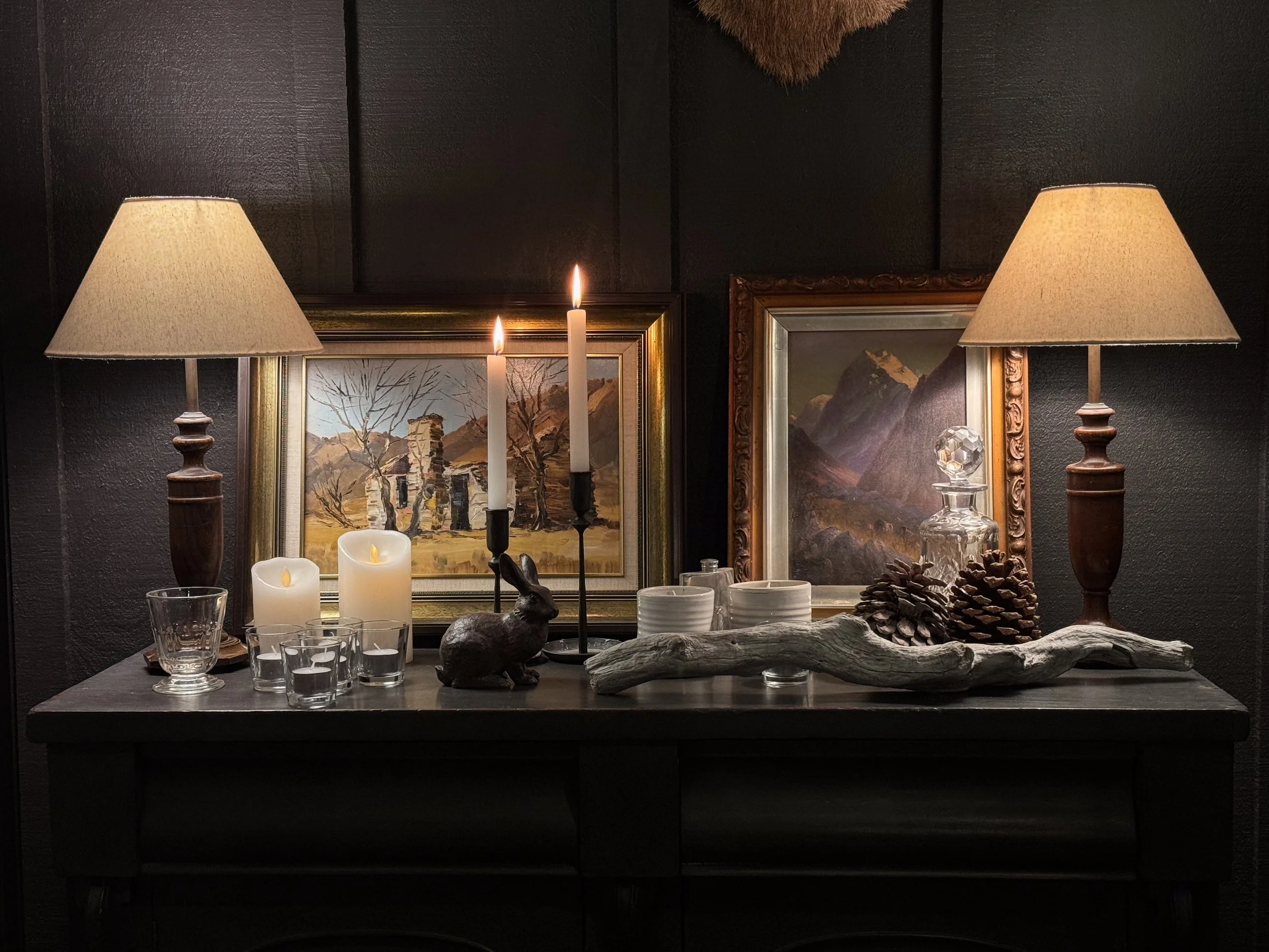A dark wooden table displays decorative candles, a ceramic bunny figurine, pinecones, and driftwood, with framed landscape paintings and table lamps with beige shades in the background.