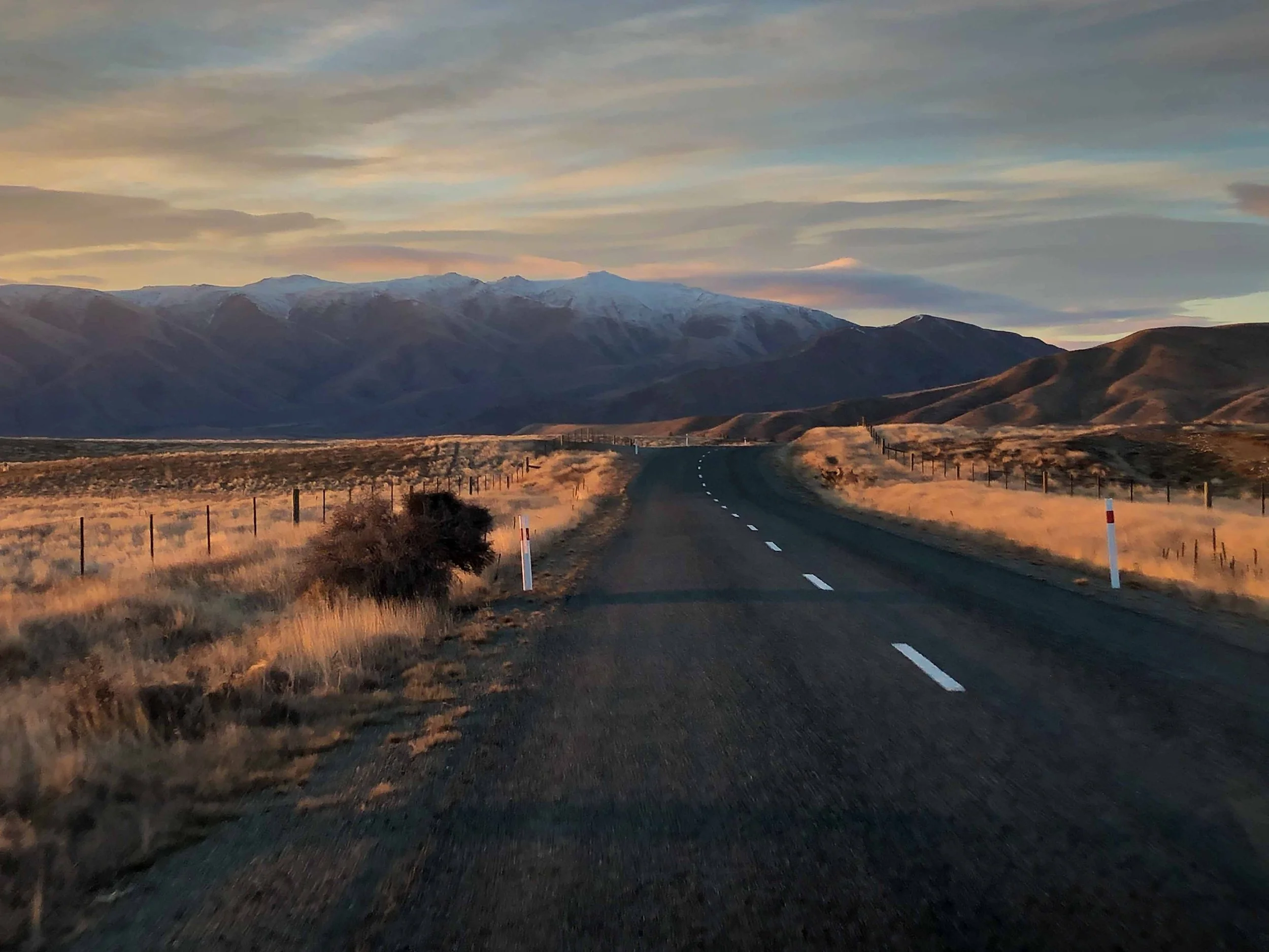 A winding two-lane road cutting through open plains with barbed wire fences on both sides, leading towards distant snow-capped mountains under a cloudy sky during sunset.