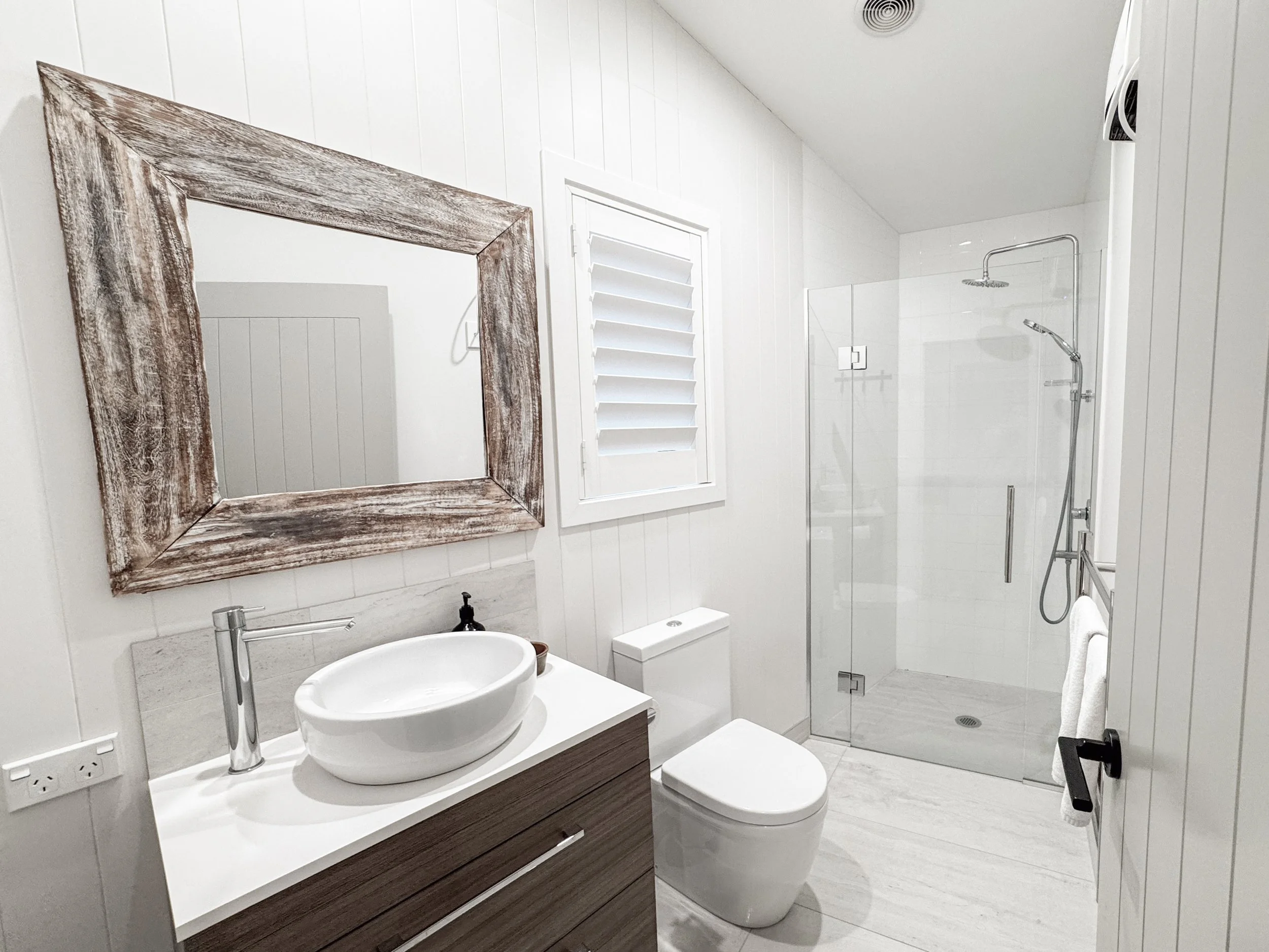 A modern bathroom with a white toilet and a bathroom vanity with a round vessel sink, a wooden framed mirror, and a gray countertop. There is a shower with a glass door and a showerhead on the far wall, and a small window with white shutters on the w