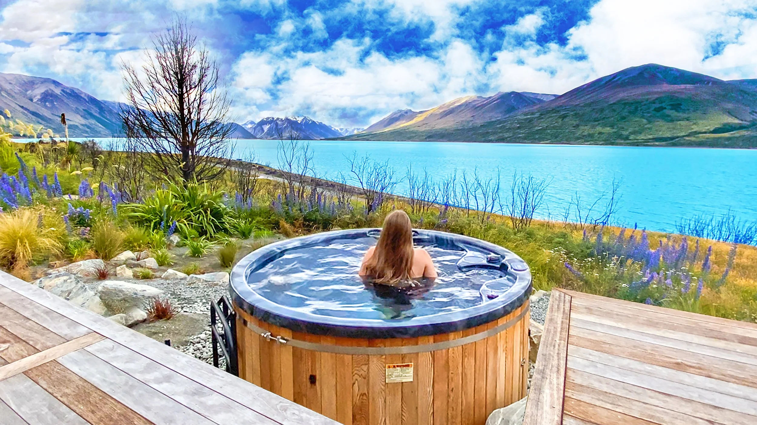 A woman with long hair relaxing in an outdoor hot tub overlooking a lake surrounded by mountains and a partly cloudy sky. Lake Ohau Escape Lake-front AirBNB Accomodation, South Island, New Zealand.