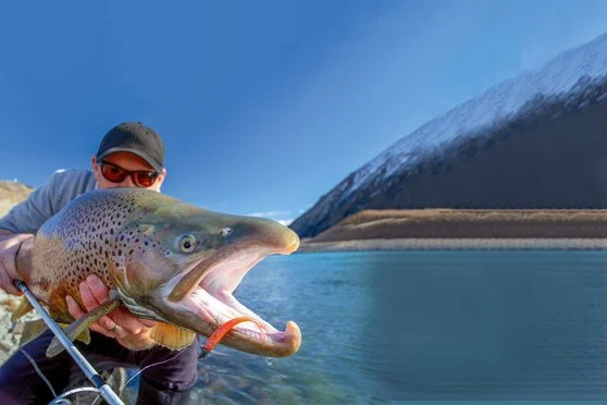 Person holding a large fish near a body of water with mountains in the background. Twizel Ohau Canal Lake River Fishing Salmon Trout. Stay at Lake Ohau Escape AirBnb Accomodation.