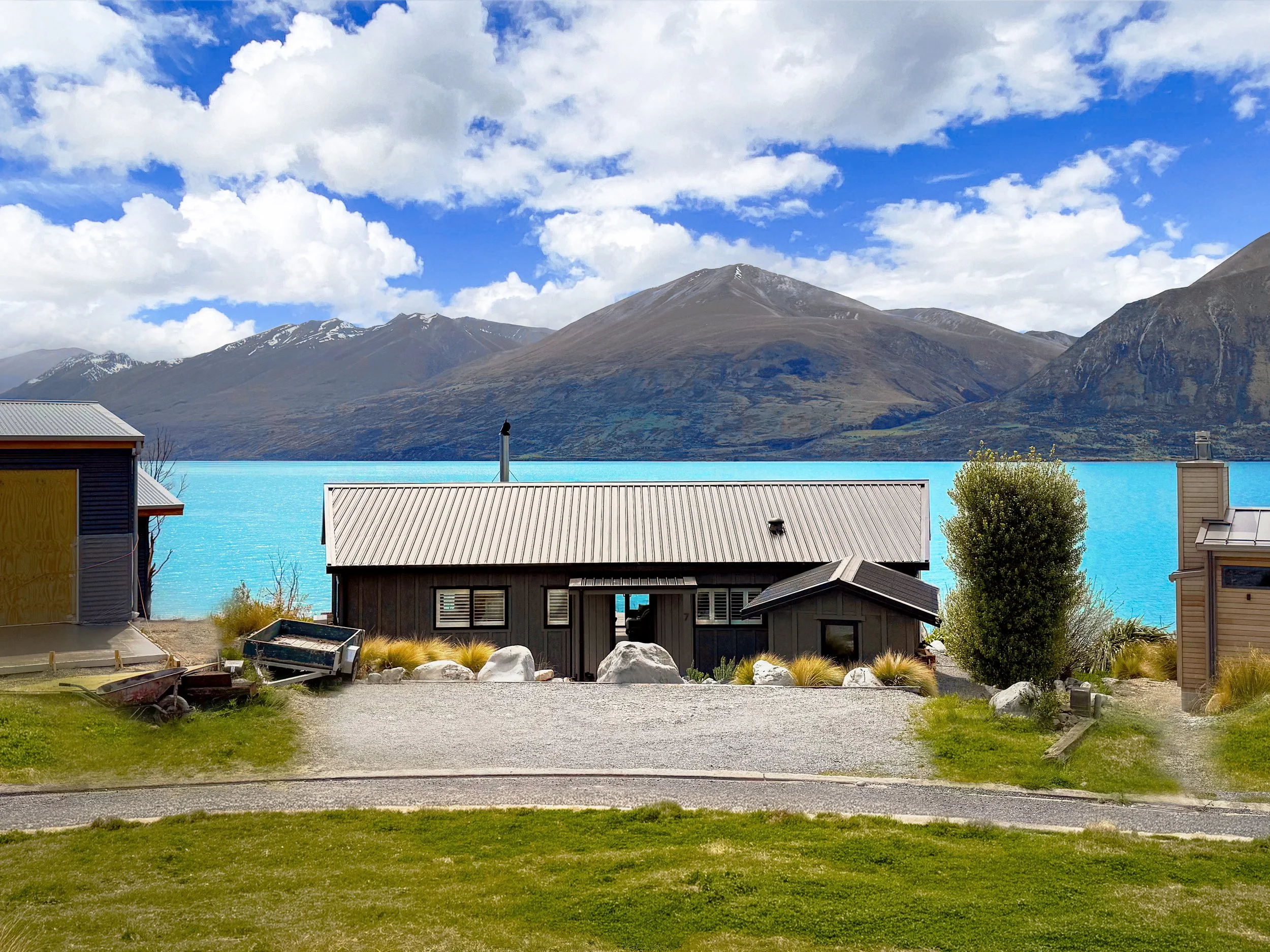 Houses with gray roofs in front of a bright turquoise lake and brown mountains under a partly cloudy sky.