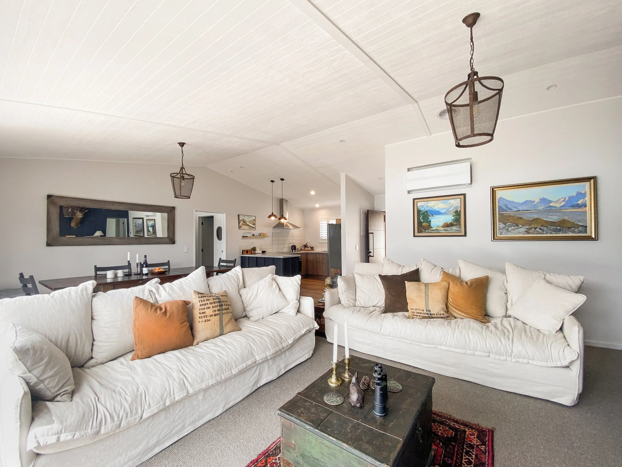 Living room with two white sofas, a black coffee table with decorative items, paintings on the wall, and a ceiling with pendant lights.