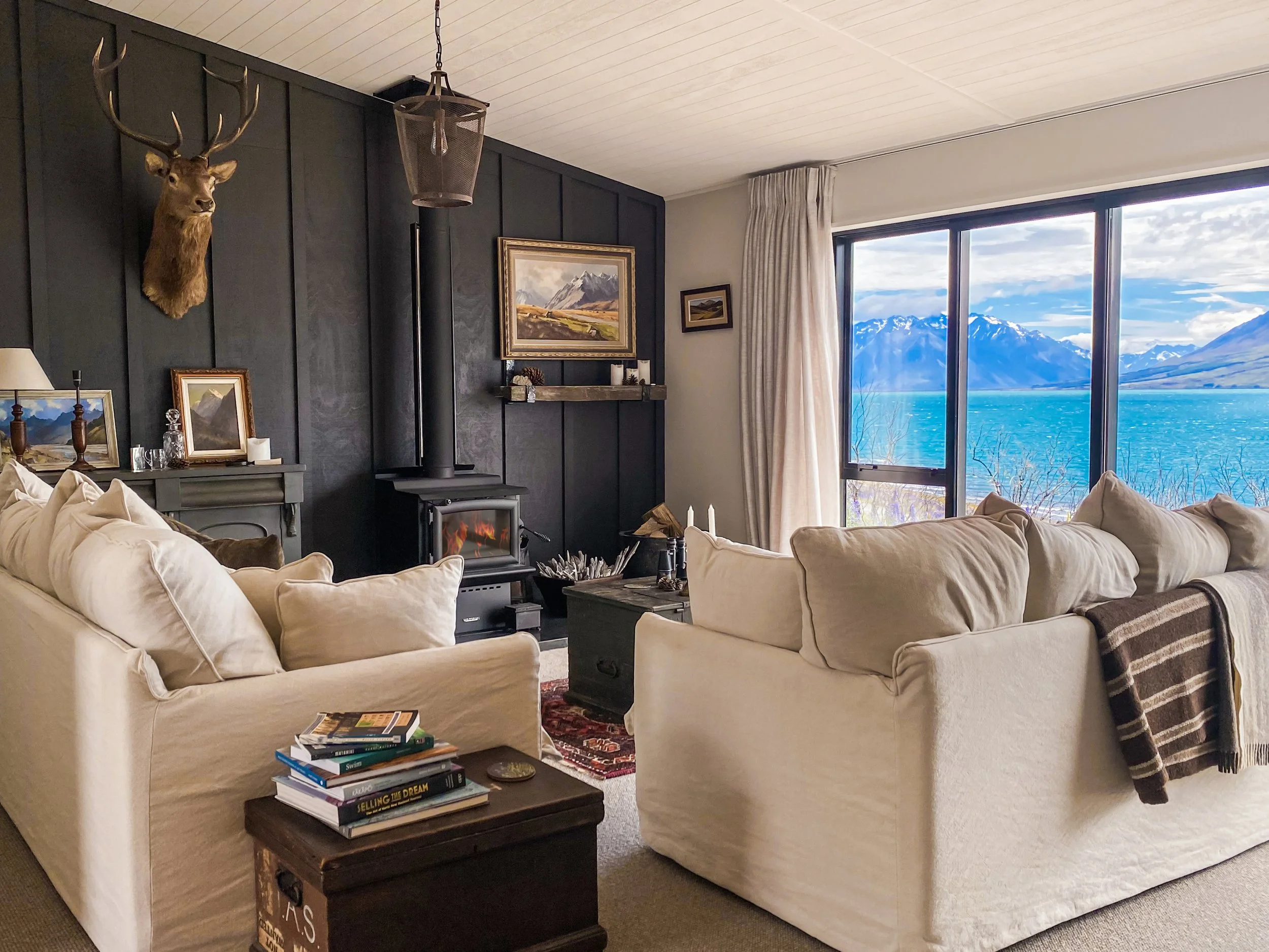 Living room with beige sofas, a dark wood table with books, a mountain and lake view outside the window, paintings on the wall, mounted deer head, fireplace, and cream curtains.