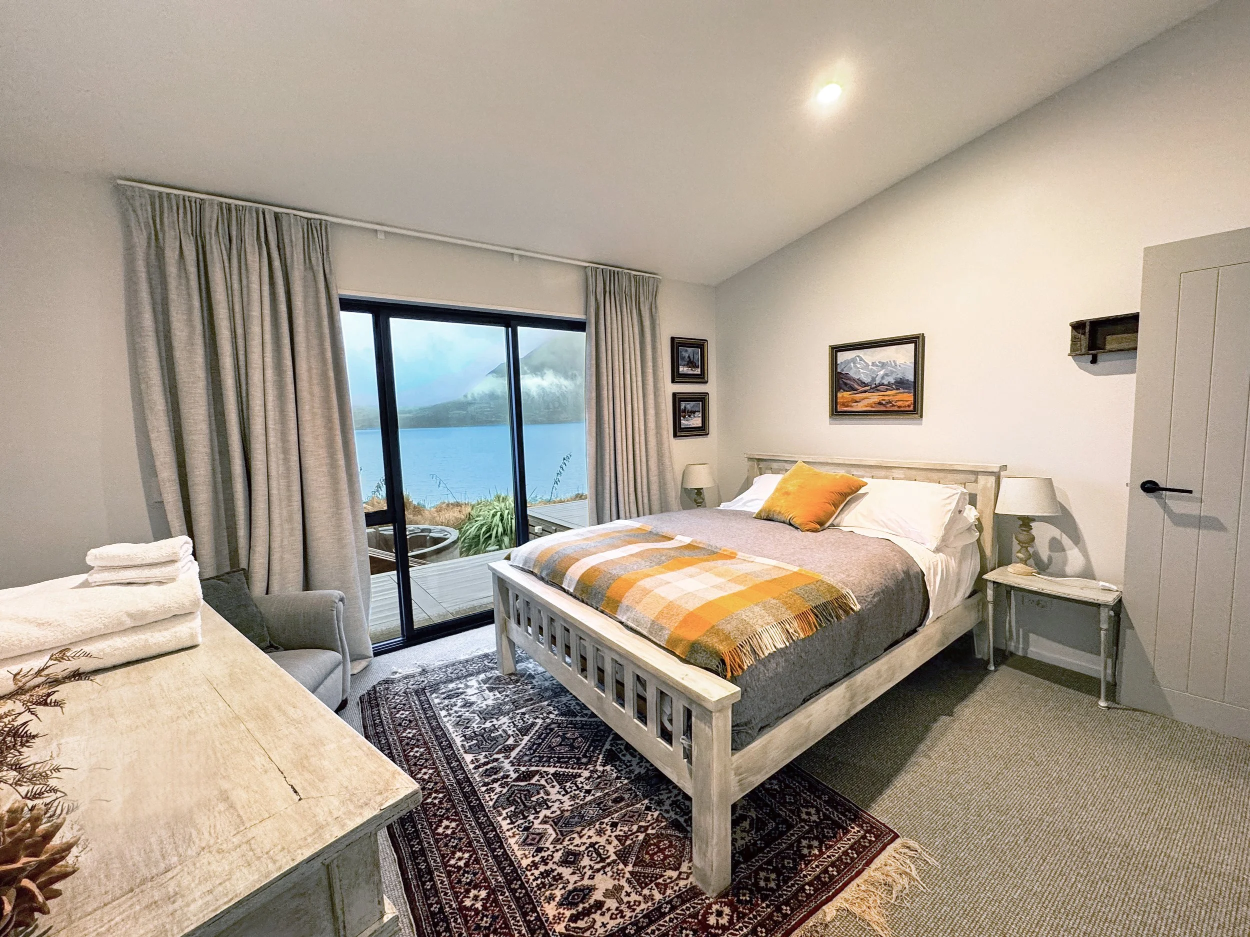 Bedroom with large window showing water and mountains, white bed with yellow and gray plaid blanket, wall art, side tables with lamps, and a sliding glass door leading to a patio with outdoor furniture.