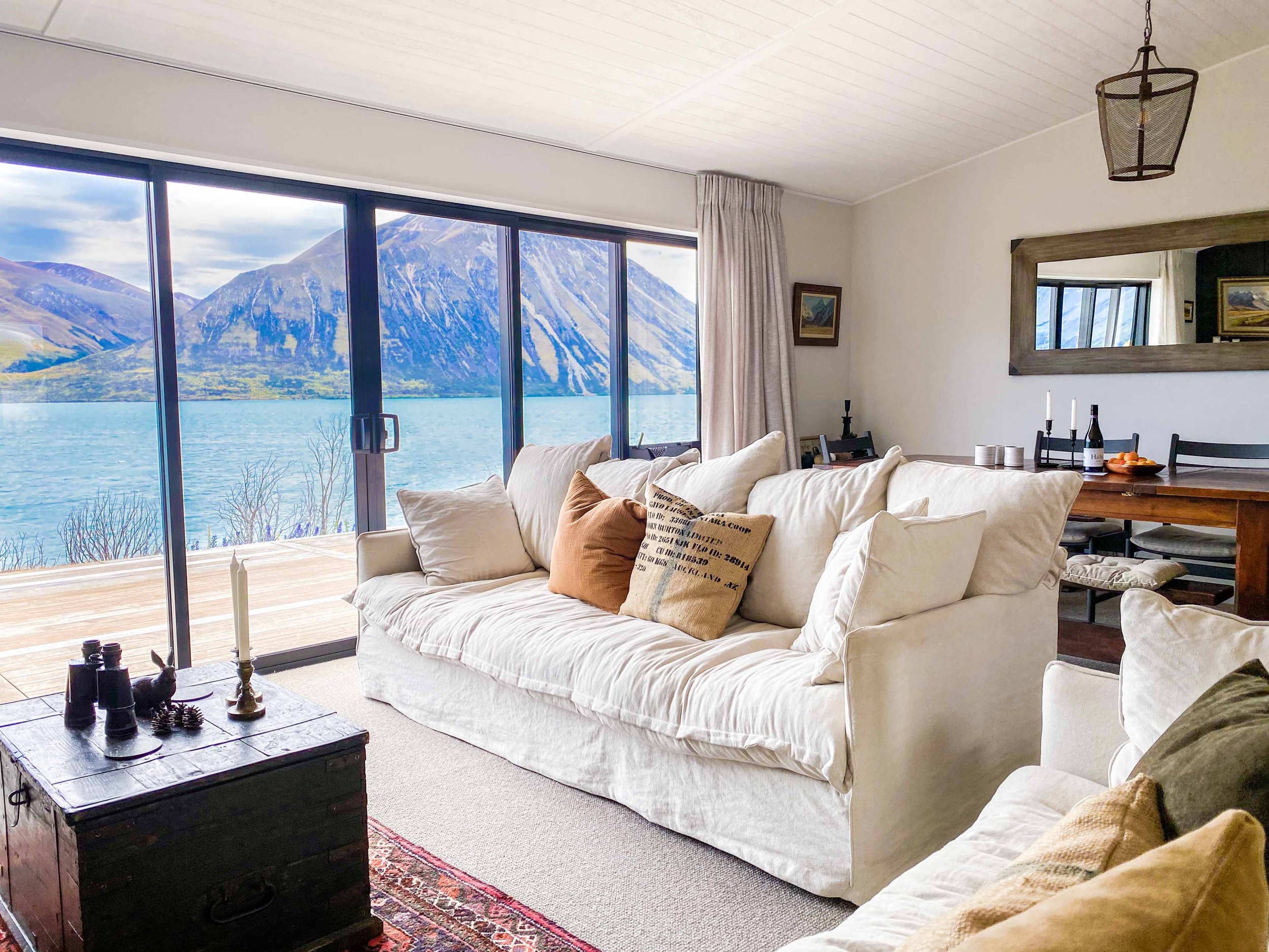 Living room with large windows showing a mountain and lake view, beige sofa with pillows, wooden coffee table with candles and binoculars, wall mirror, chandelier, and dining table with wine bottles and fruit in the background.