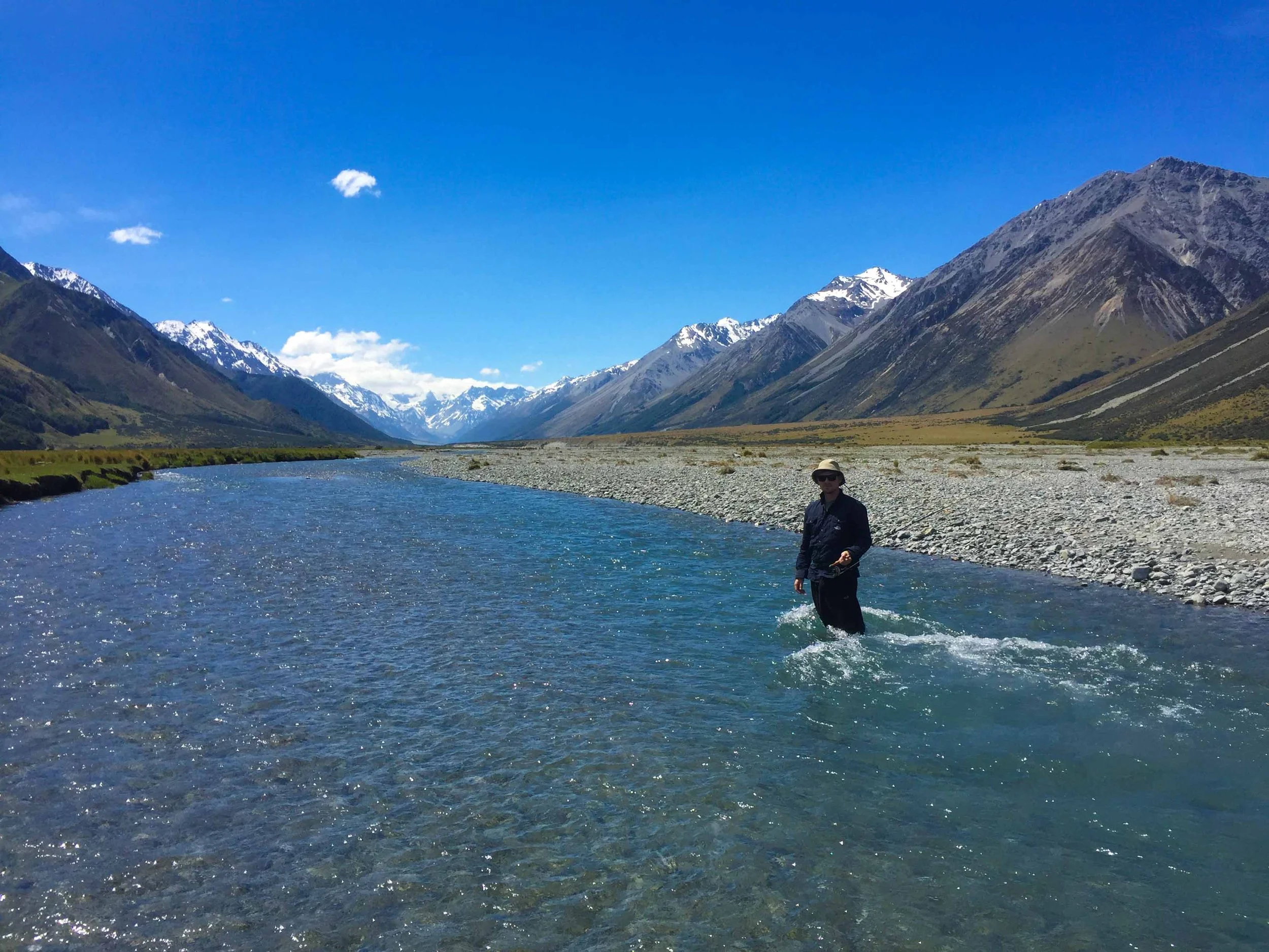 A person standing in a river with mountains in the background, snow-capped peaks under a clear blue sky.