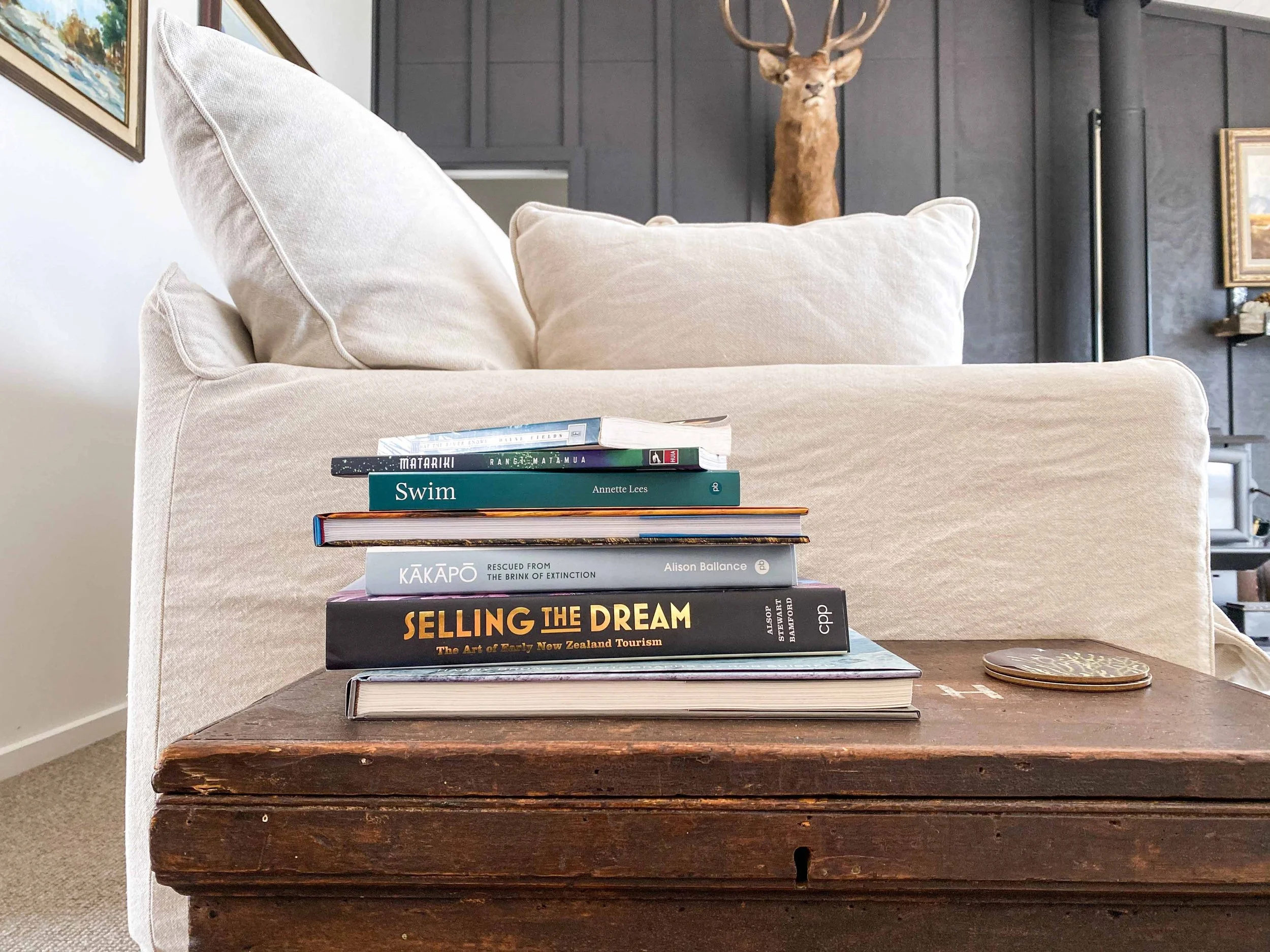 A stack of books sitting on a wooden table in front of a beige sofa, with a deer head mounted on a dark wall in the background.