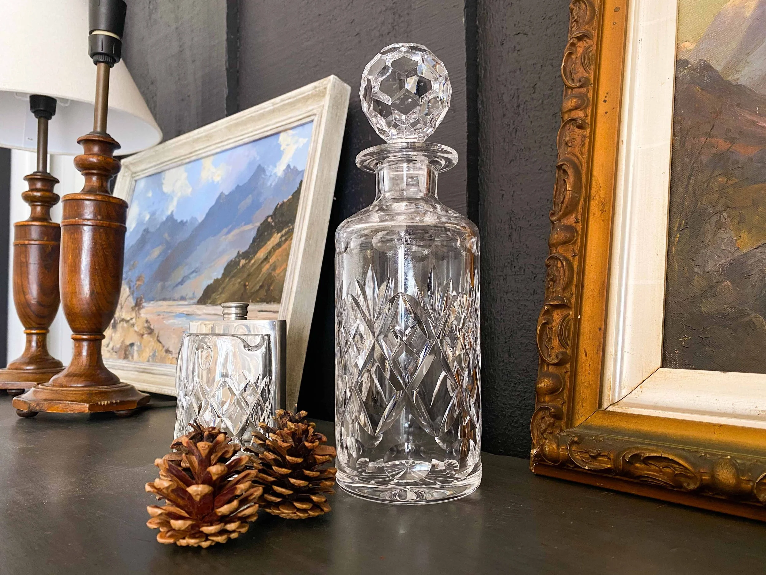 Decorative table setting featuring a crystal decanter with a diamond-shaped stopper, two pinecones, and a painting. In the background, two wooden candlesticks with black candles and a model ship are visible.