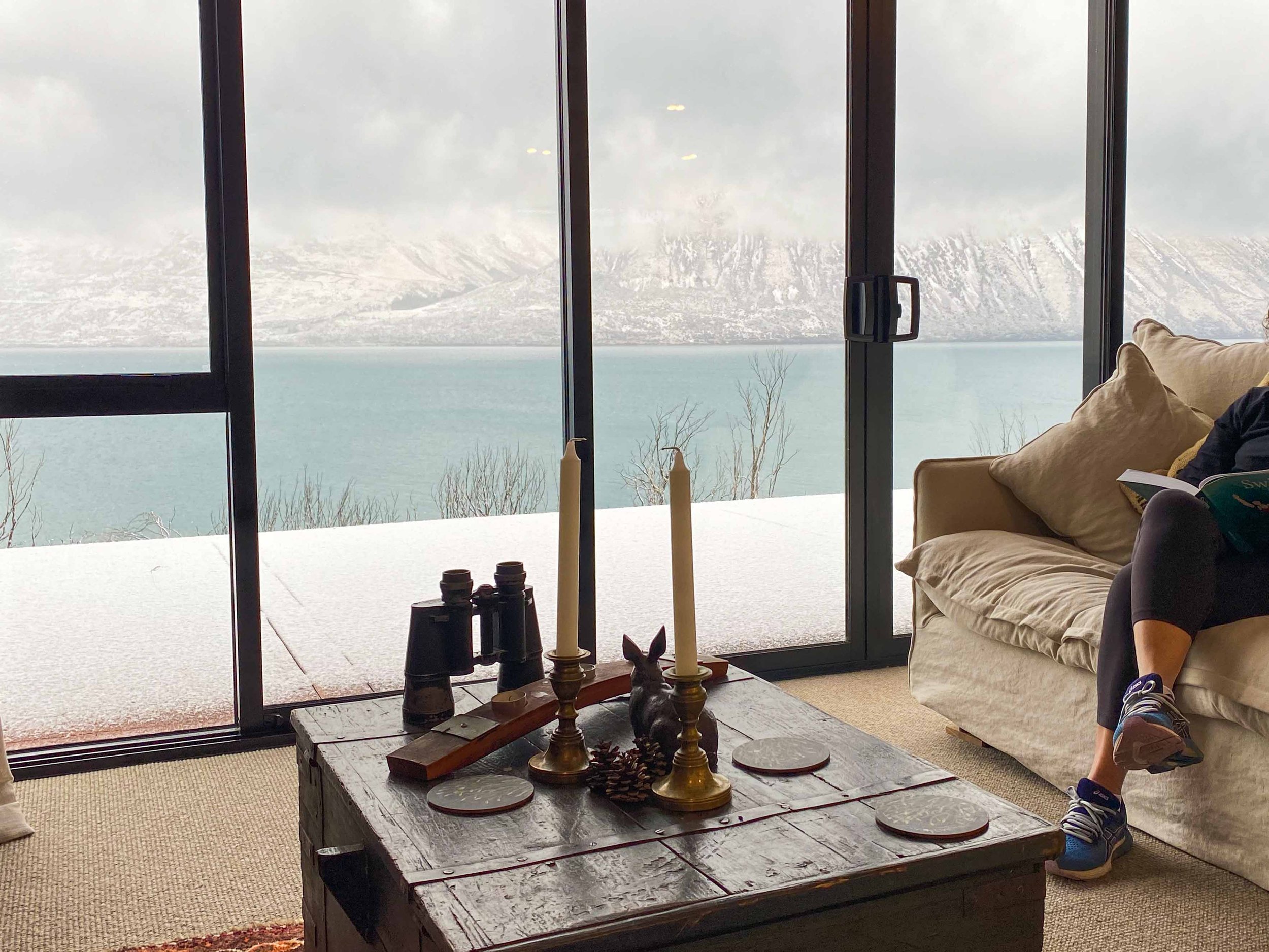 A cozy living room with a large window view of snowy mountains and a body of water. Inside, there is a wooden coffee table decorated with candles ornaments, a beige sofa with cushions, part of a person's leg and shoe visible on the right side. Ohau. 