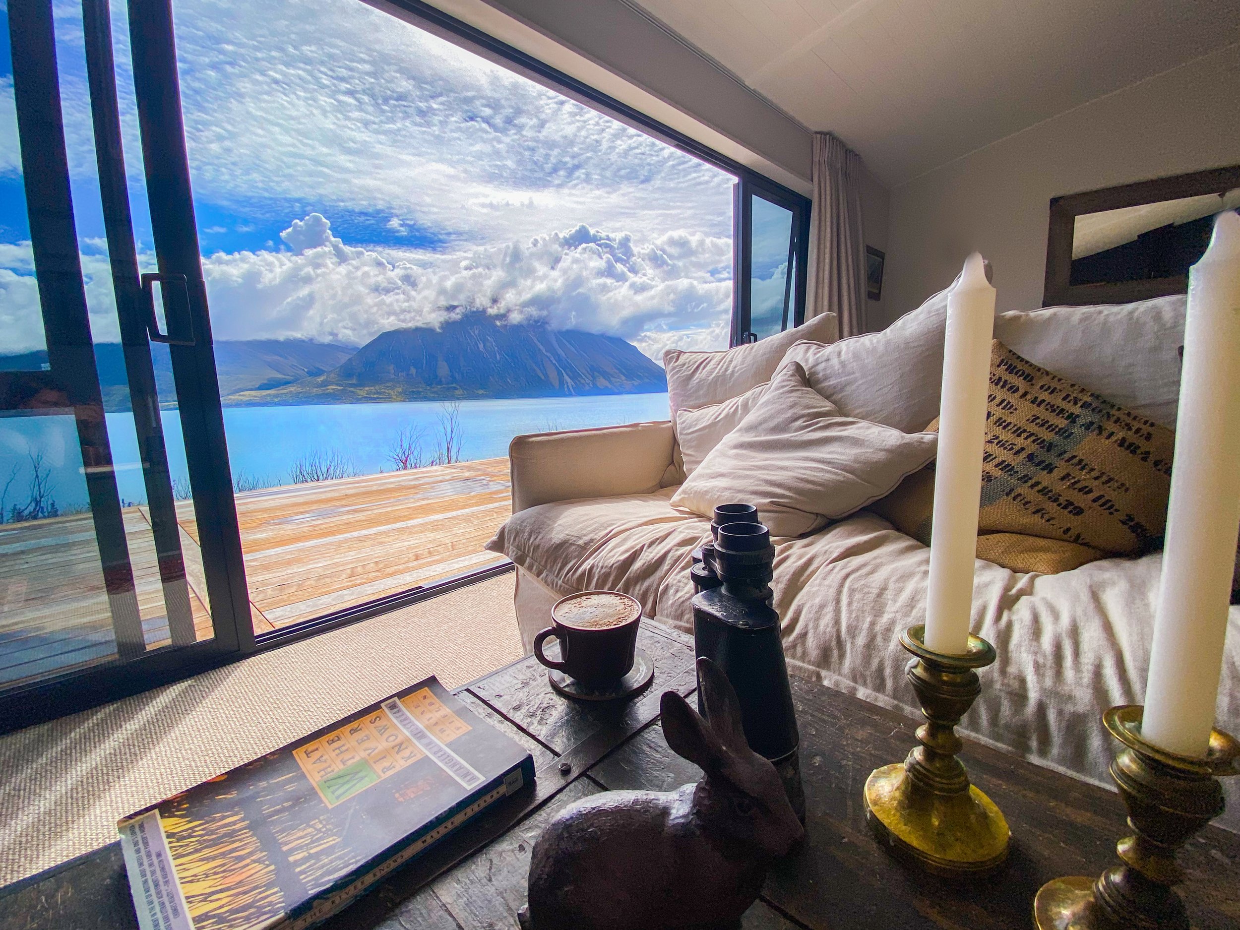A cozy indoor living space with a large open window showing a scenic view of mountains, a lake, and a cloudy sky, with a sofa, candles, coffee cups, and magazines in the foreground.