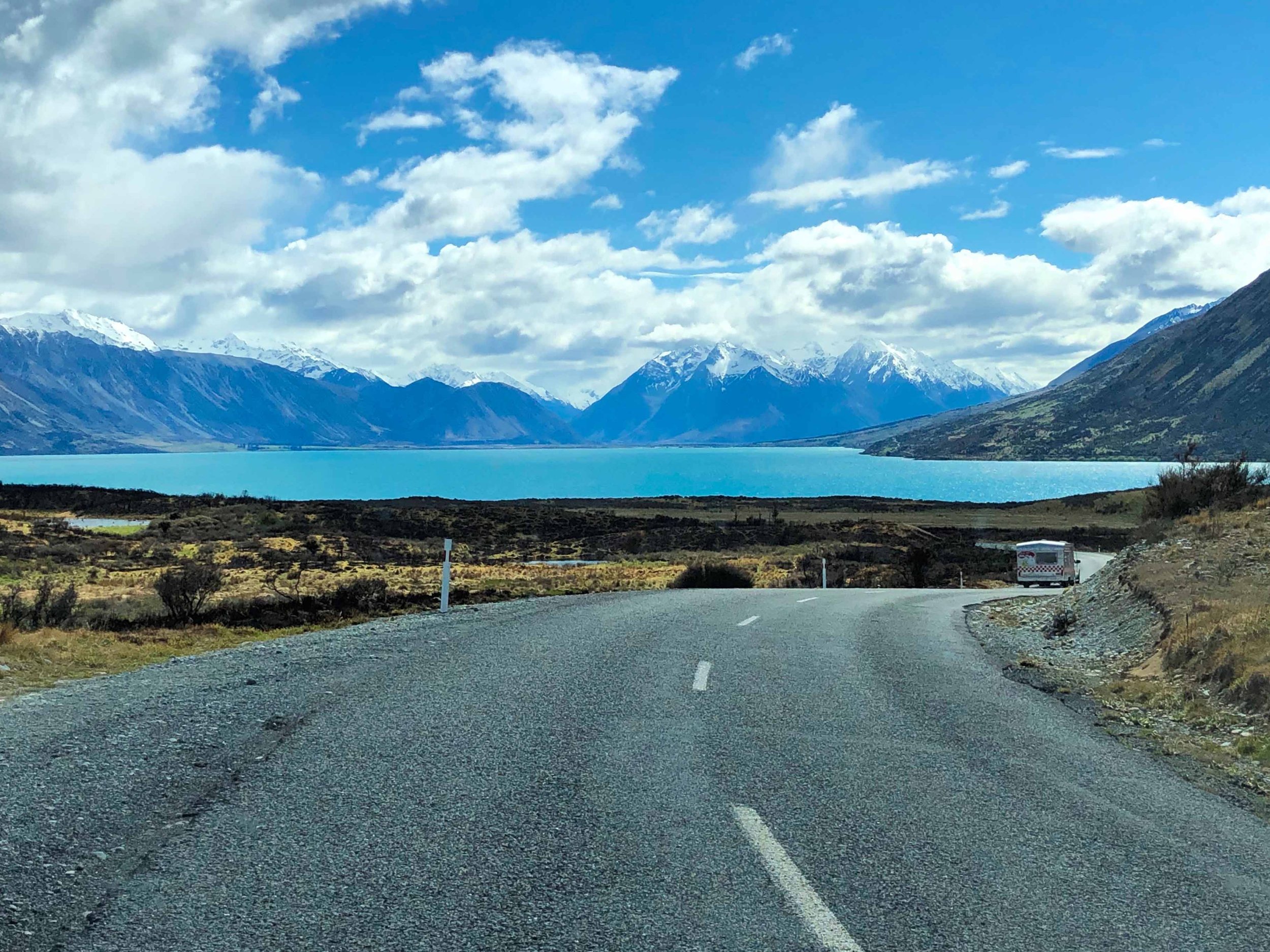 A winding road near a lake with mountains in the background, some with snow, under a partly cloudy sky.