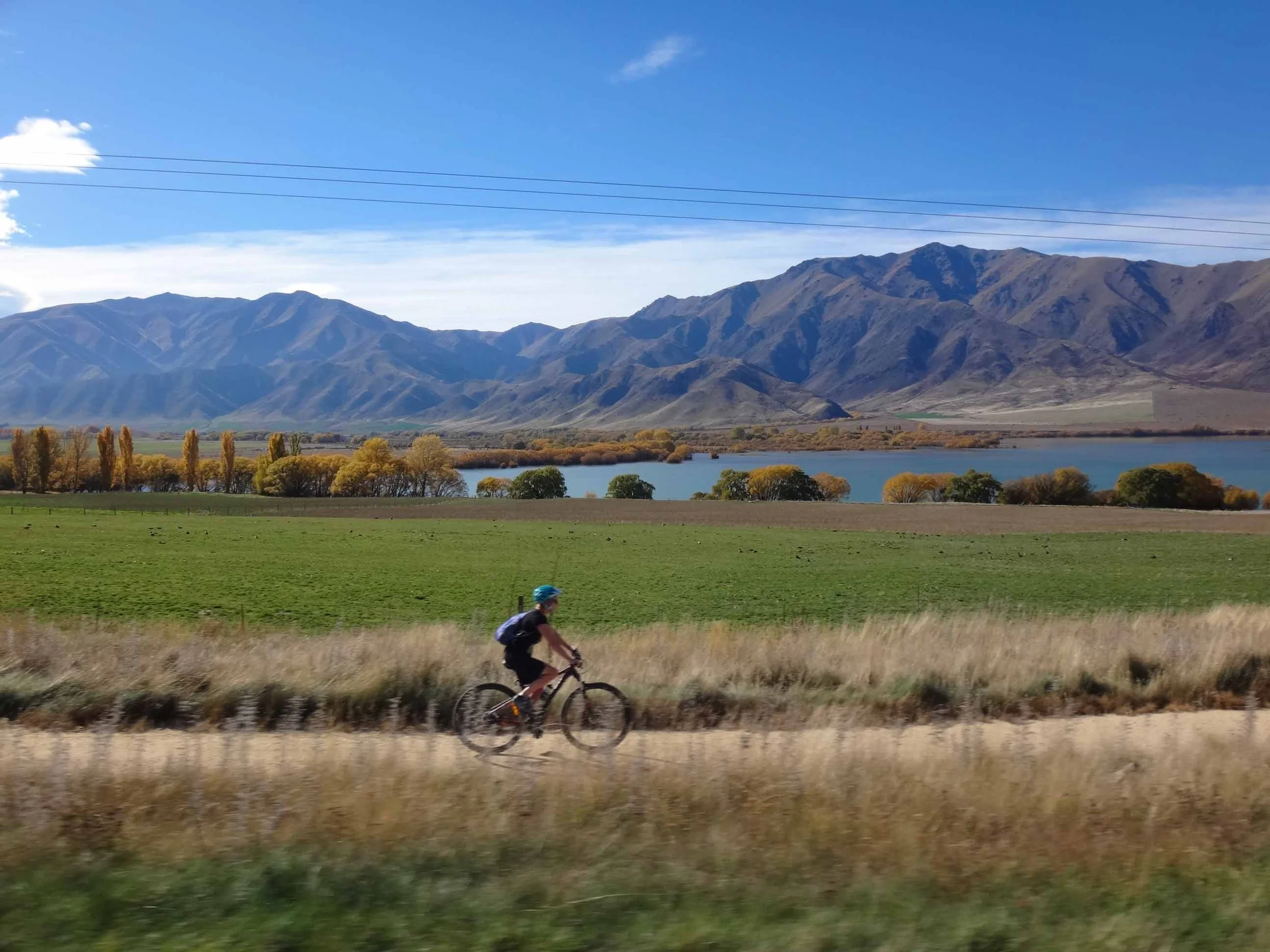 A person riding a bicycle along a rural path with mountains, trees, a lake, and a partly cloudy sky in the background.