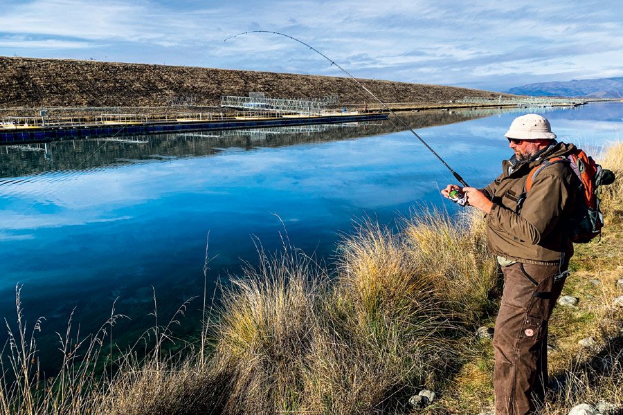 A man fishing by a calm river with mountains and a dam in the background, wearing a hat, sunglasses, a brown jacket, and carrying a backpack. Twizel Ohau Canal Lake River Fishing Salmon Trout. Stay at Lake Ohau Escape AirBnb Accomodation.