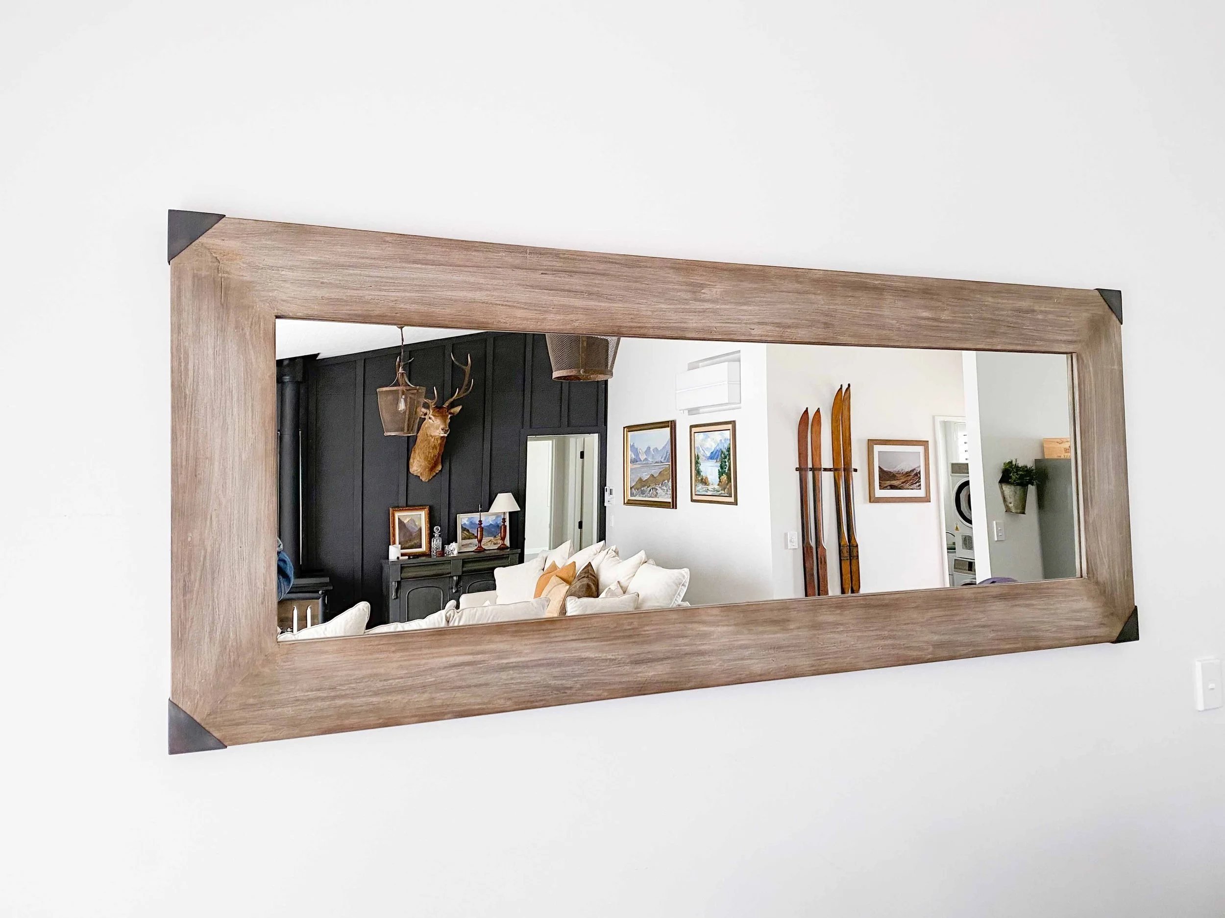 A large rectangular mirror with a wooden frame hangs on a white wall, reflecting a living room with framed artwork, a dark cabinet, and a mounted deer head.